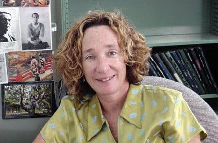 Headshot of poet Jane Miller in a yellow shirt with polka dots, sitting in front of a book shelf.