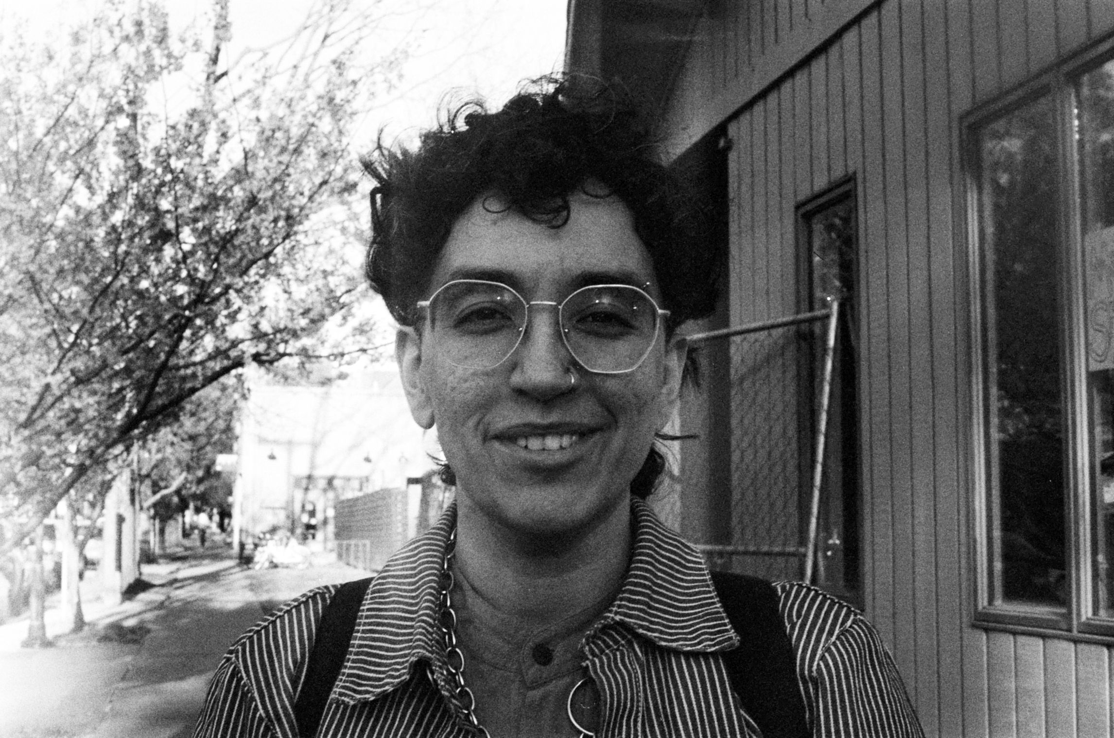 This is a black and white photo of Aishvarya Arora, a South Asian person with short, black curly hair, seen from the shoulders up, wearing a striped shirt and a thick chain necklace standing on a sidewalk with trees in the background.