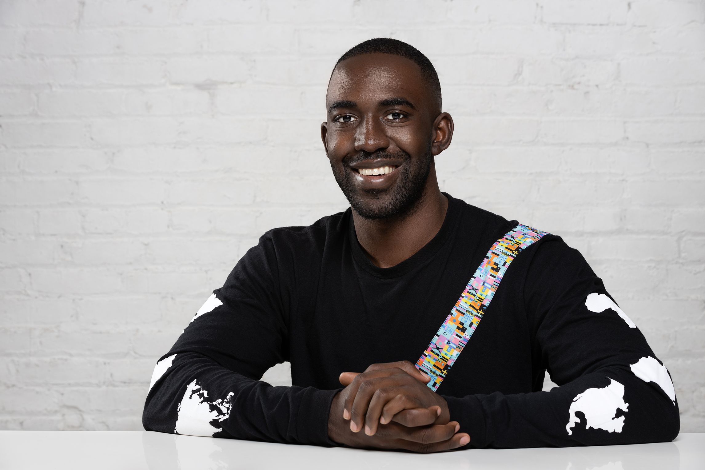 Bryan Byrdlong, a Black man with short black hair and a black long sleeve shirt sits in front of a white brick wall