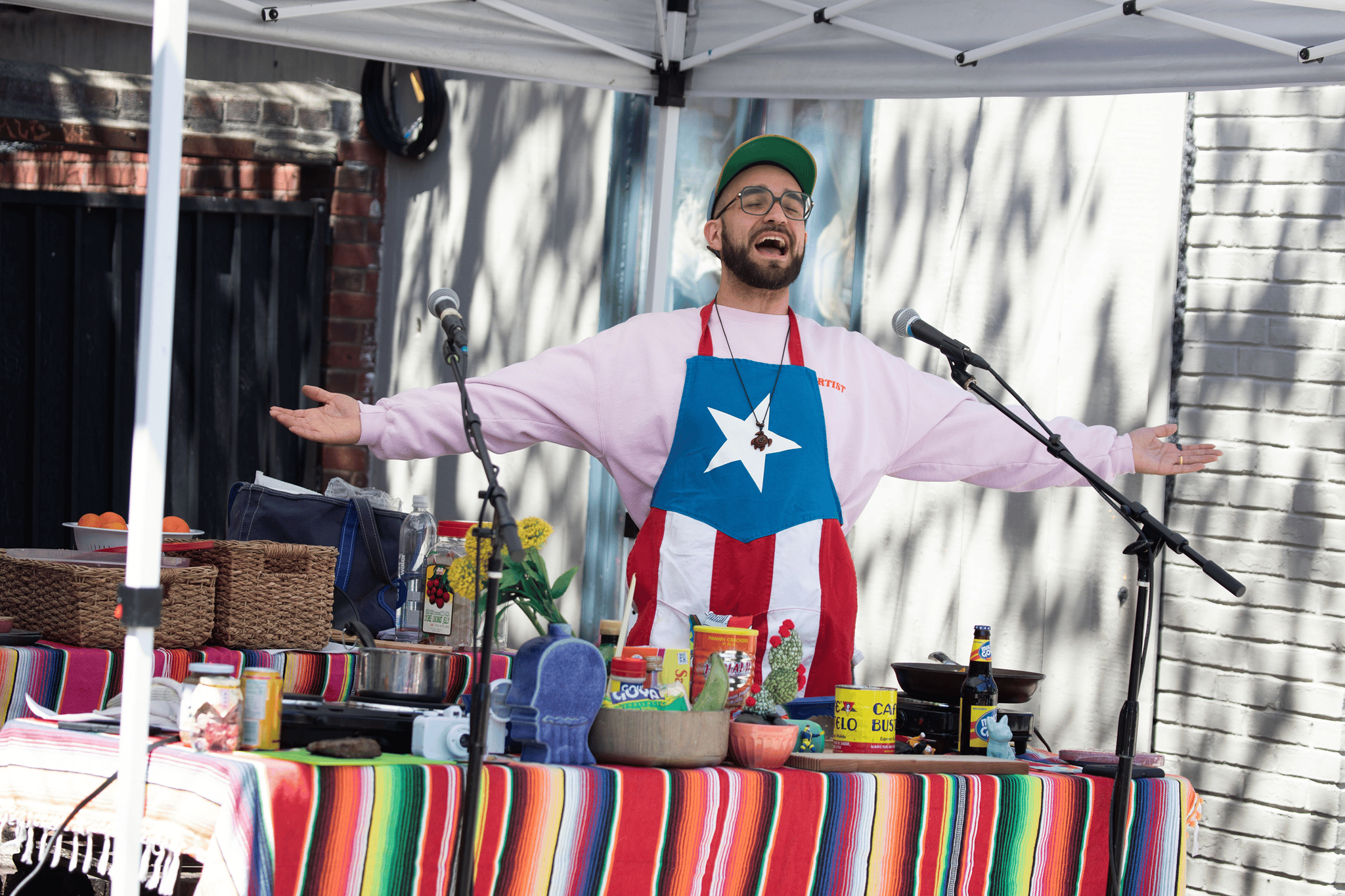 Man standing at a booth covered in colorful blankets. His arms are outstretched, his mouth is open to speak, and his eyes are closed. He's wearing an apron of the Puerto Rican flag.