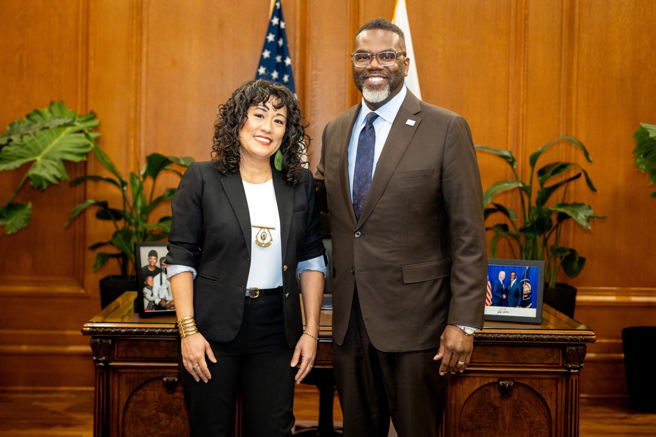 Mayda del Valle and Chicago Mayor Brandon Johnson standing and smiling in a wood paneled office with two flags and plants behind them.