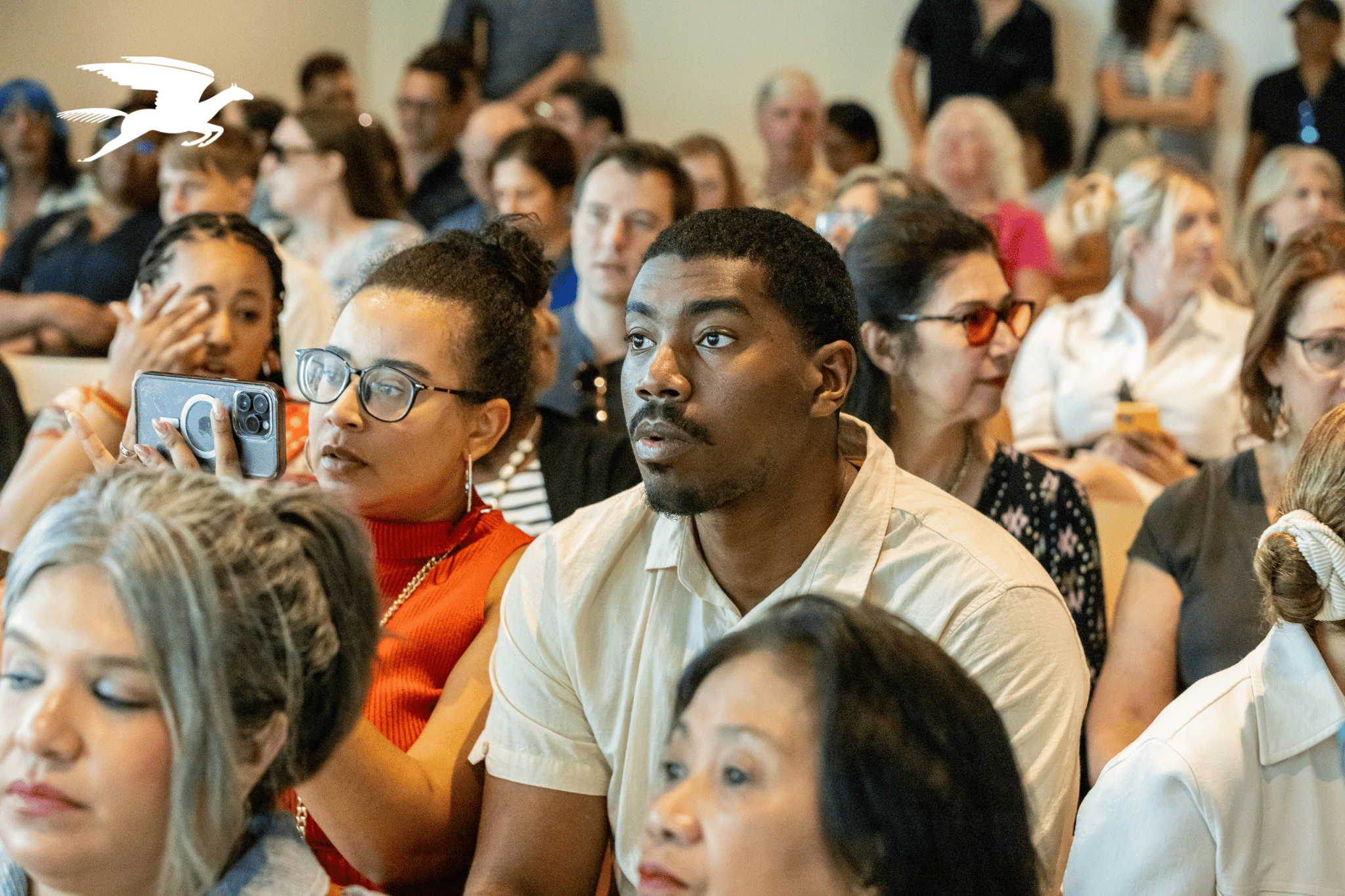 Room of adults in chairs looking forward raptly. A white pegasus logo appears in the upper left.