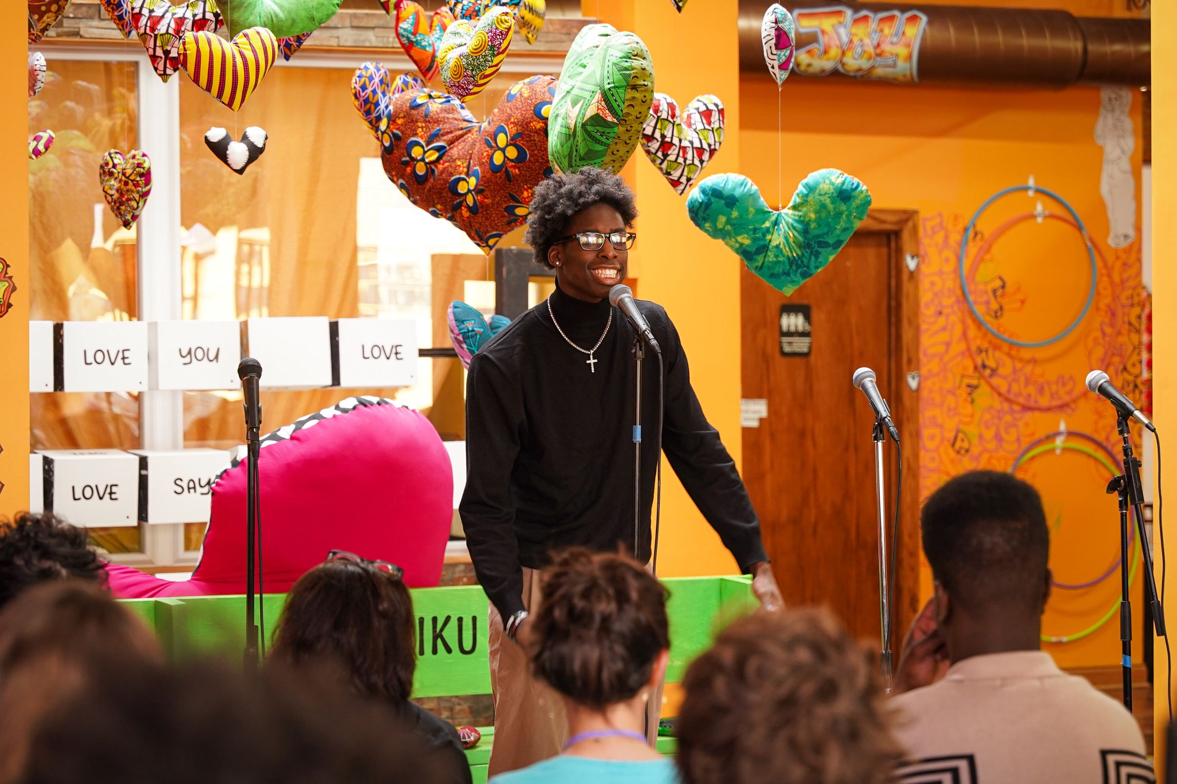 Young person at a microphone in front of a seated group in a colorful room with balloons floating behind him