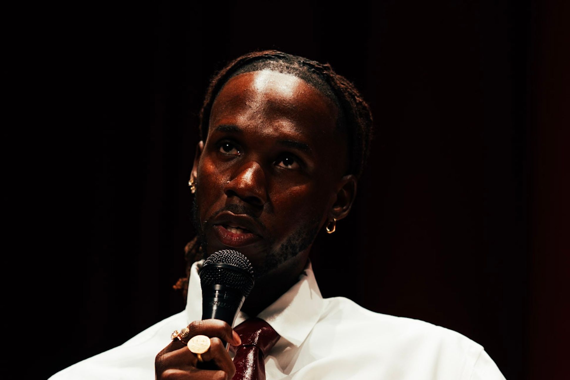 W.J. Lofton, a Black man with locs tied back, wearing a white shirt and red leather tie, holds a microphone while seated.