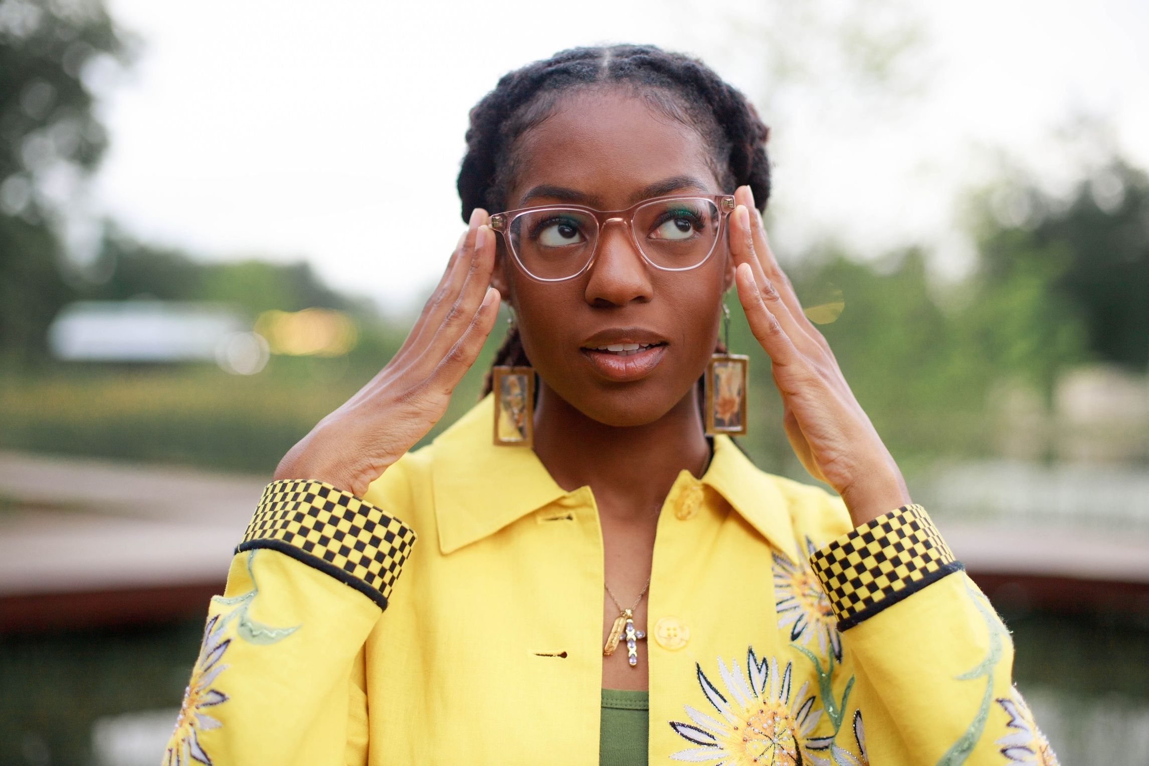 Aris Kian, Black woman with locs wearing yellow jacket with sunflowers holding her hands to her glasses.