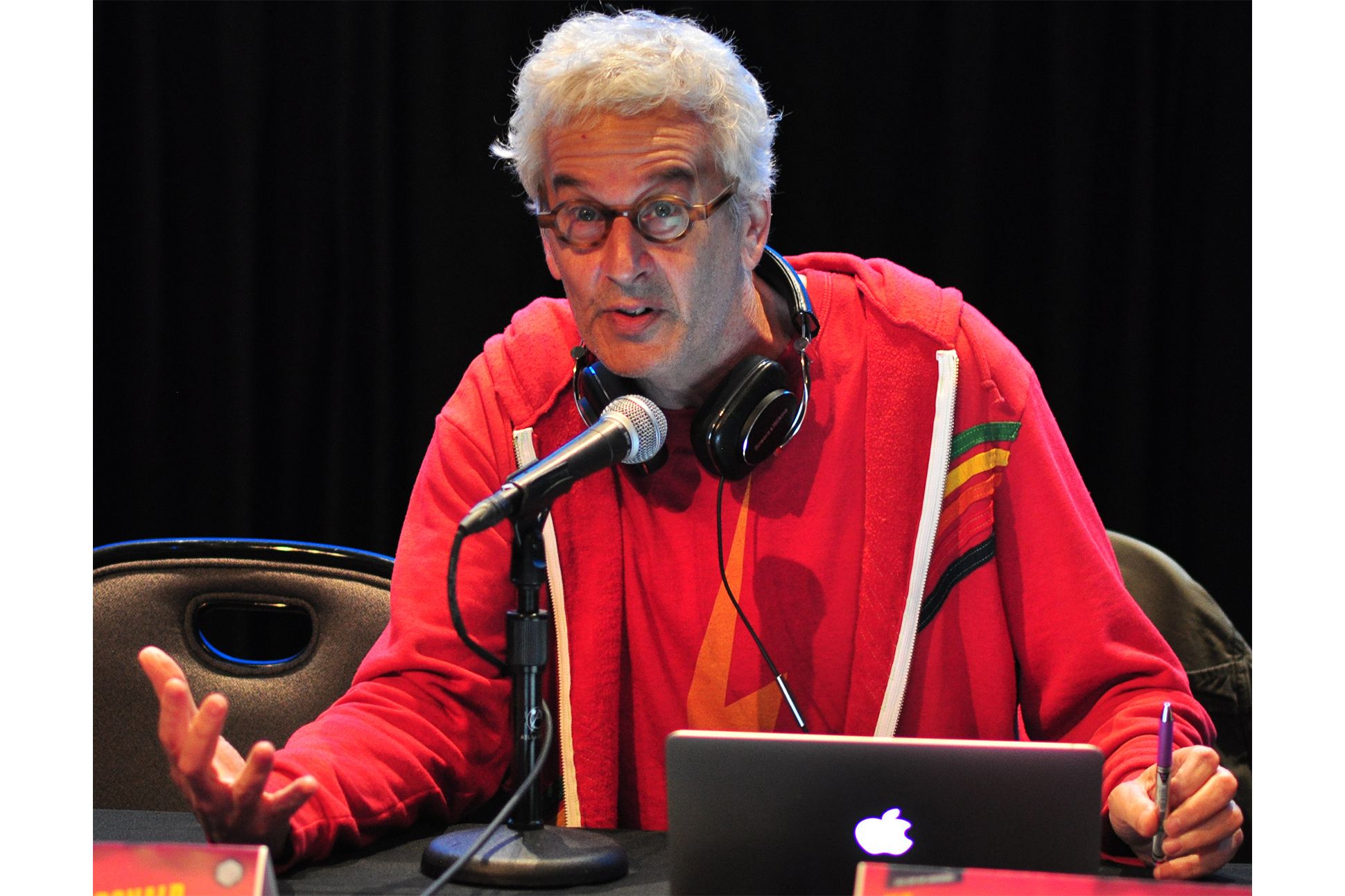 Male with white hair, wearing a red top and brown-rimmed glasses sits at a conference table with a mike in front of home and headphones around his neck, gesturing with his right hand while holding a pen in his left hand.