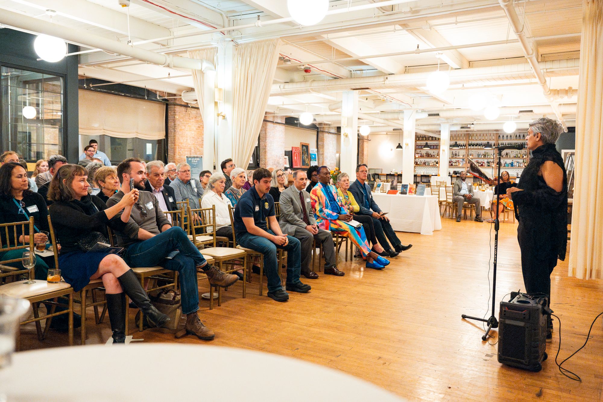 Room of adults seated in chairs while an adult stands at a microphone in front of them