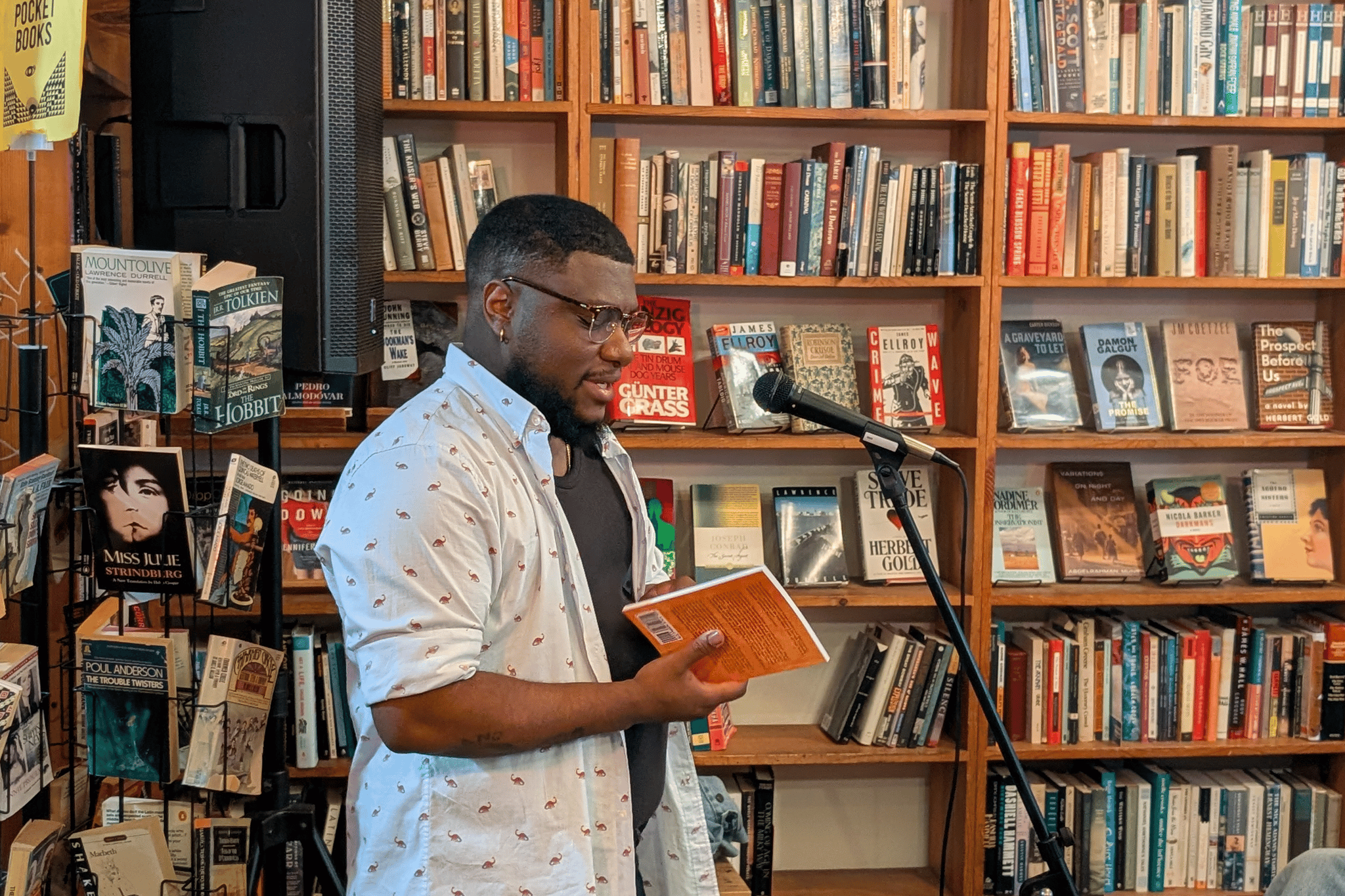 Adult standing at a microphone holding an orange book. They are surrounded by bookshelves.