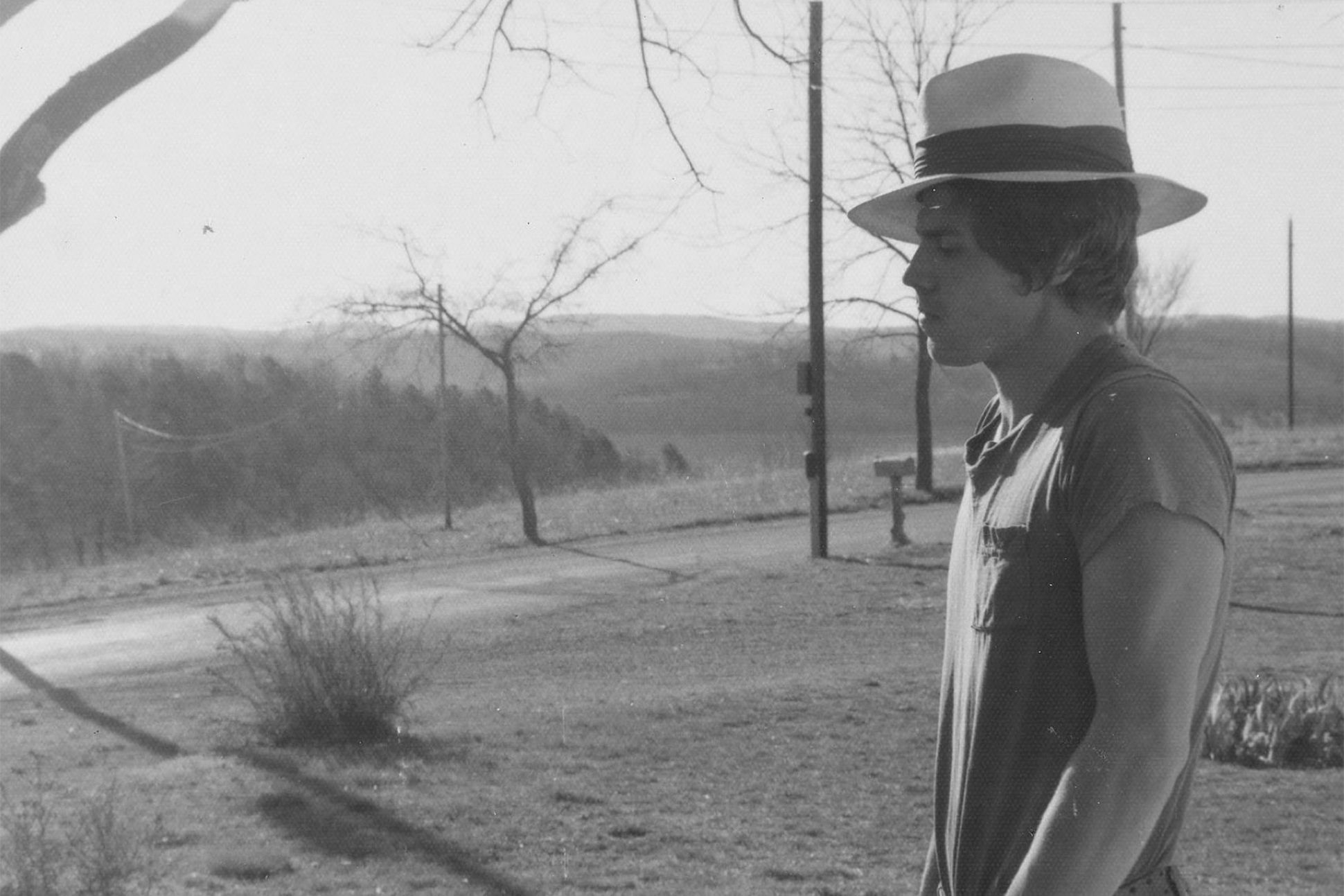 A black-and-white photograph of Frank Stanford wearing a hat and a t-shirt, standing outside in a rural environment.