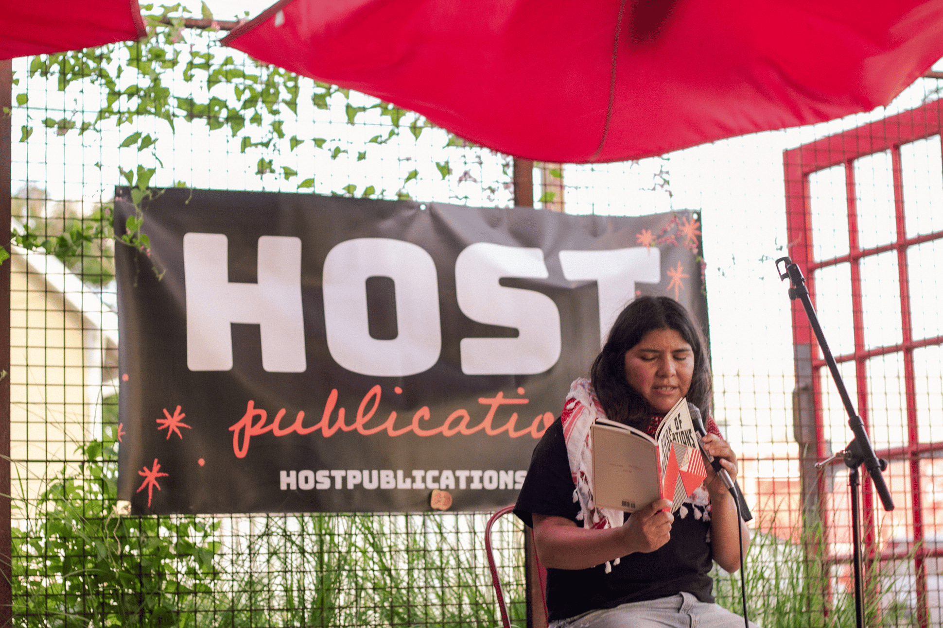 mónica teresa ortiz reading a book on a stage under a red canopy outdoors with a black and red Host Publications banner in the background.