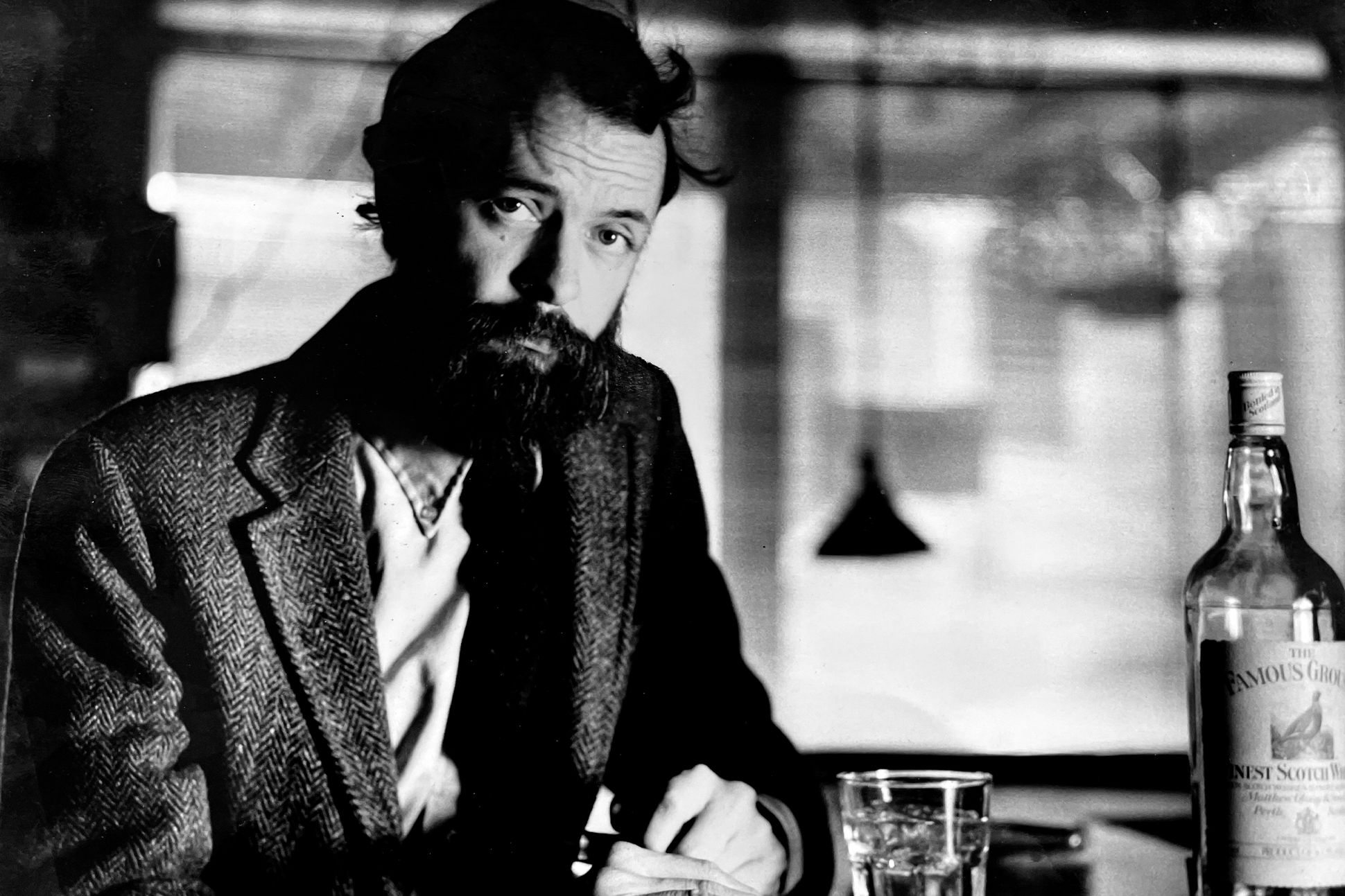 A black-and-white photograph of Everette Maddox sitting at a bar with a shot glass and a bottle of alcohol nearby.
