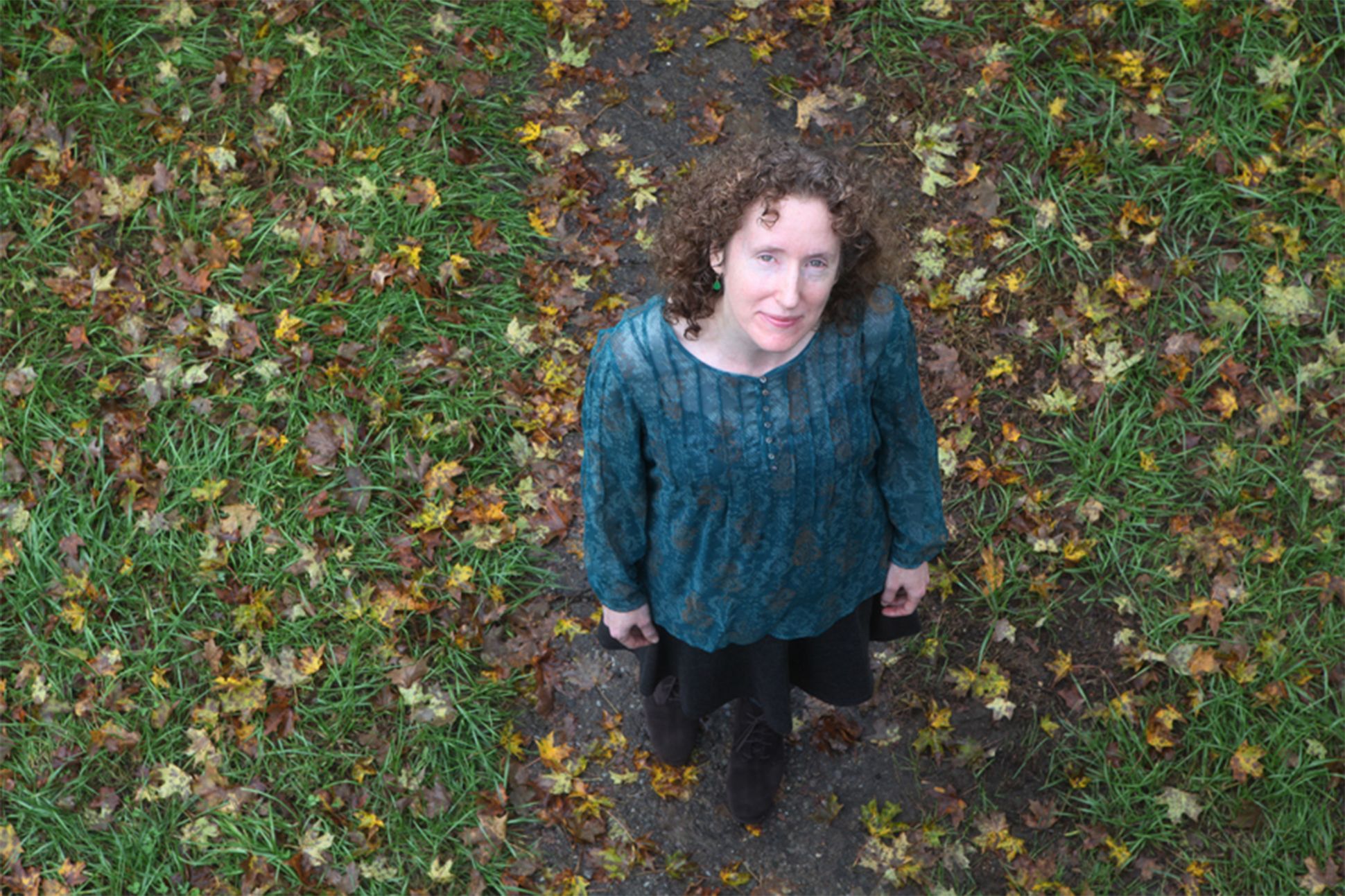 Photo of Charlotte. Mandell, shot from above, in a field of fallen green and brownish leaves. Mandell is gazing up at the camera, her face framed by curly hair, wearing a turquoise top.