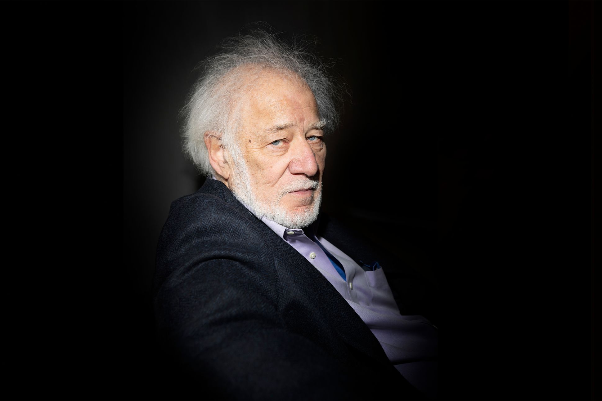 A photograph of Michael Ondaatje looking into the camera, spotlit amid darkness.