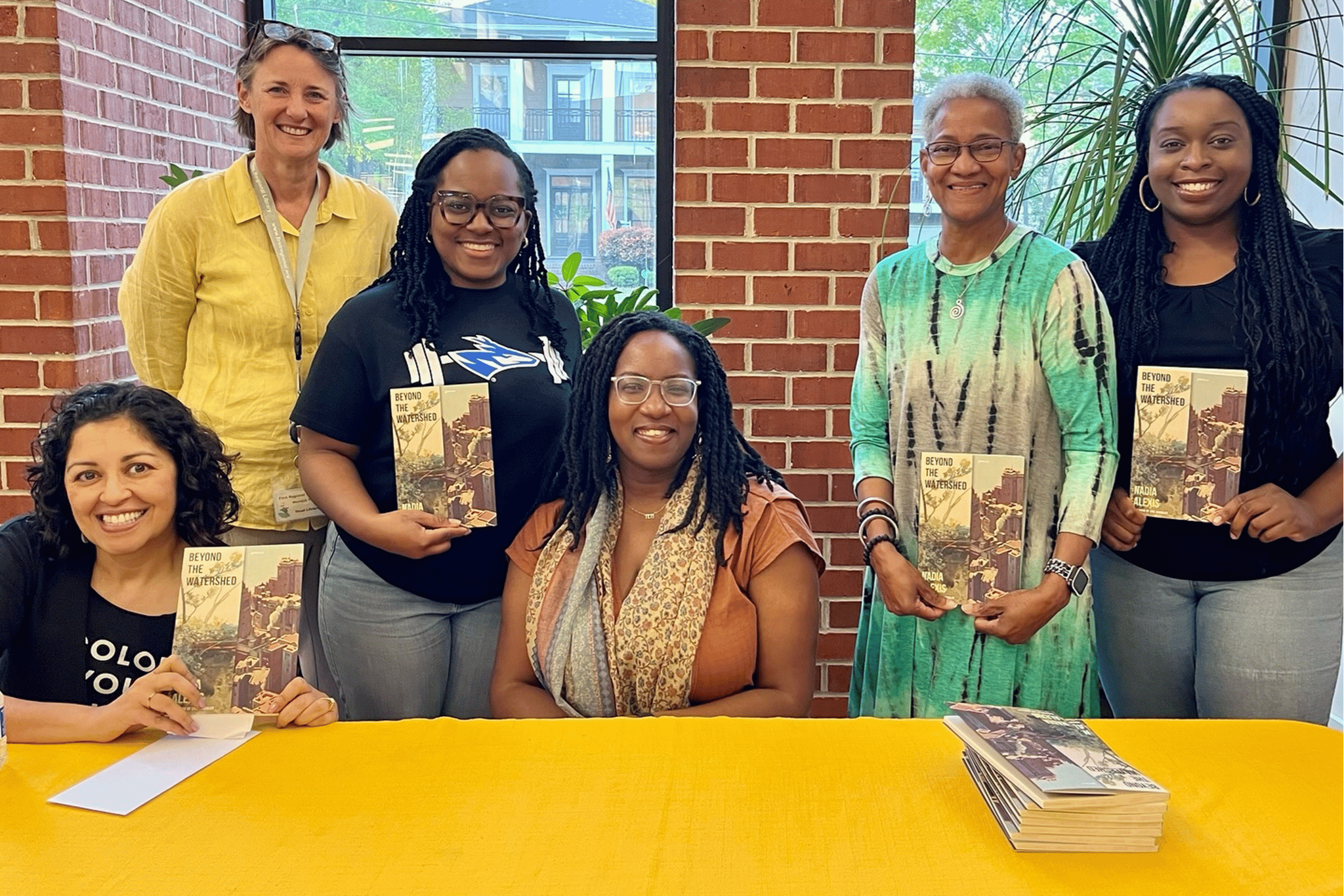 Six adult women sitting and standing behind a yellow table holding copies of the book Beyond the Watershed,
