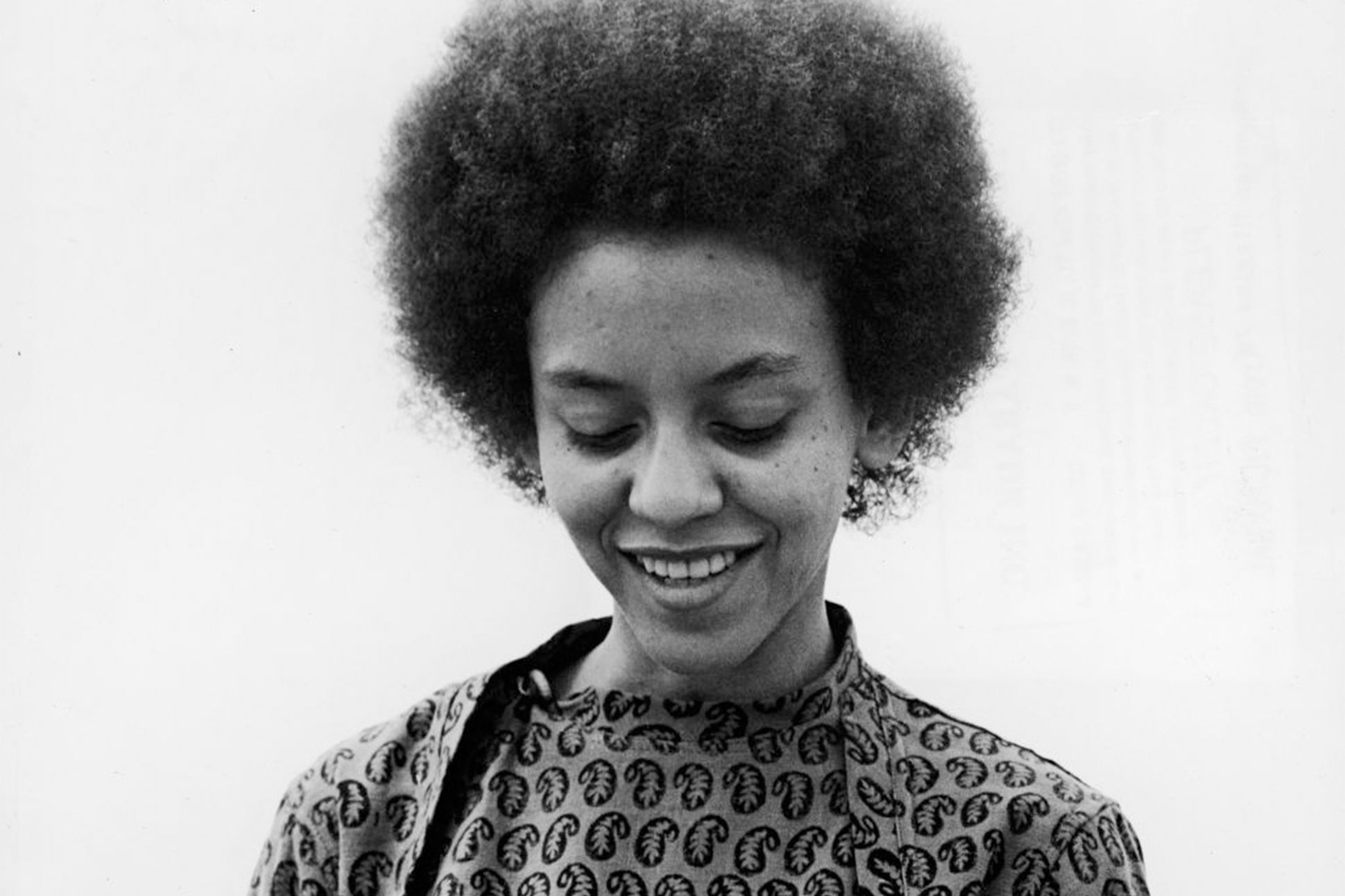 A black-and-white photograph of Nikki Giovanni smiling and looking down.