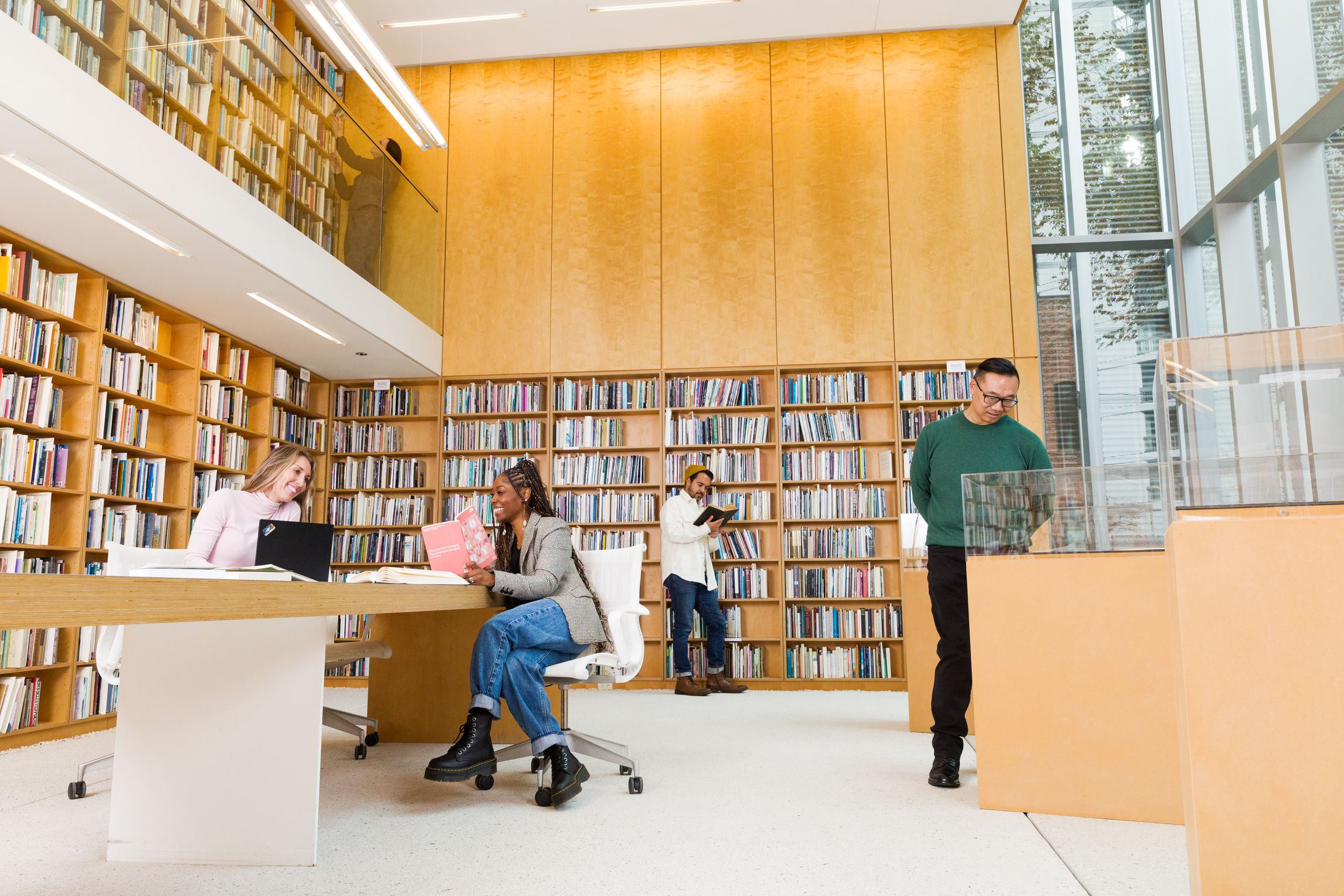 Two-story room with light wood panelling and bookshelves filled with books. On one side are two stories of windows. Two adults are seated at a table with a laptop and a book. One is standing with a book, and another is standing at a display case.