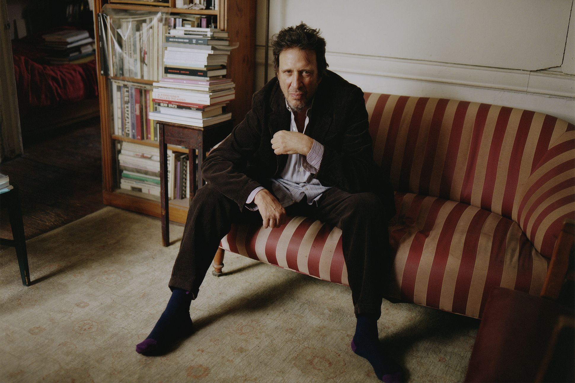 A photograph of Richard Hell sitting on a striped couch, looking into the camera.
