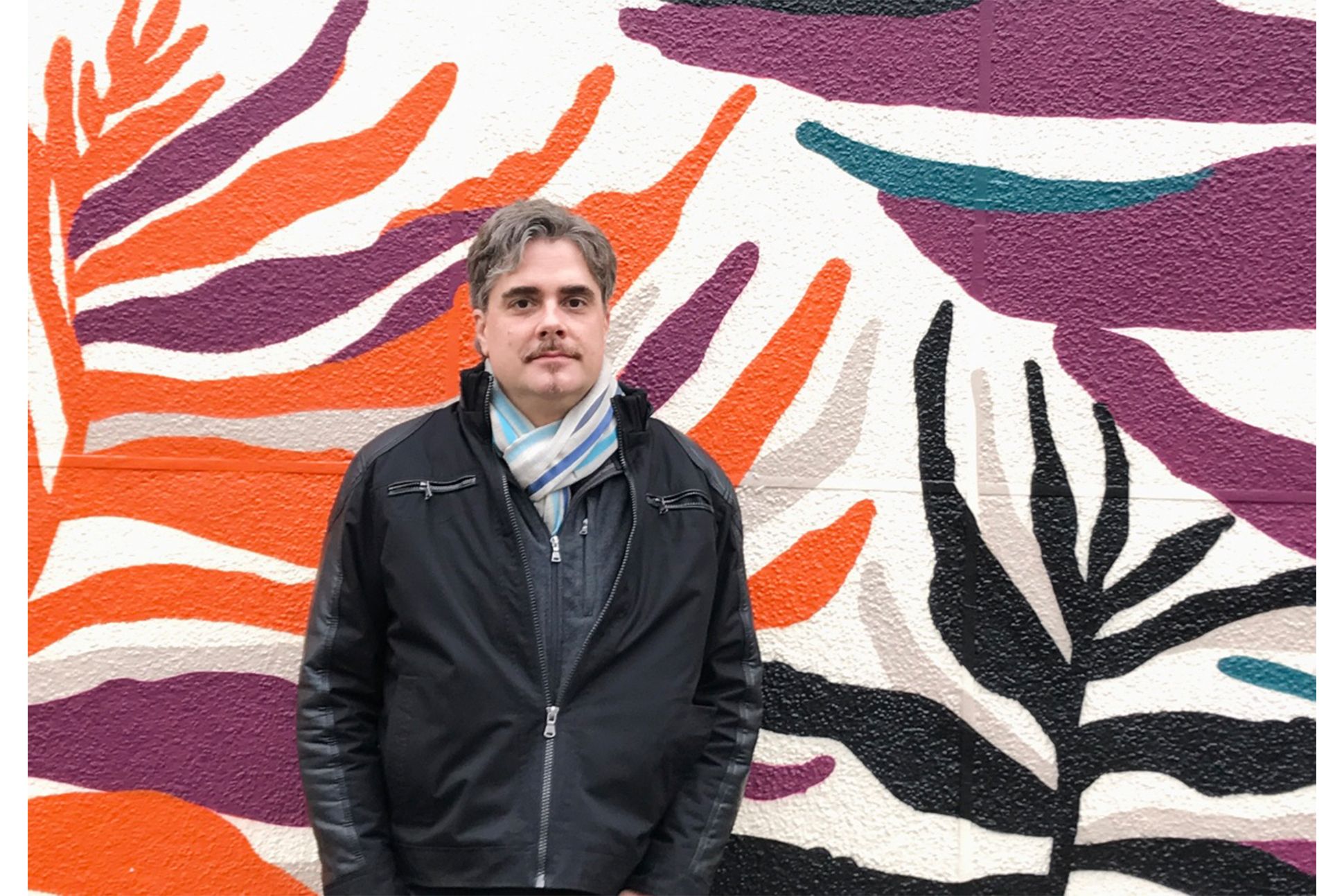White male with short greyish hair wearing black jacket and a white scarf with blue lines, standing in front of a wall mural featuring organic, plant-like shapes in purple, orange, black, cream, and turquoise, against a white background.