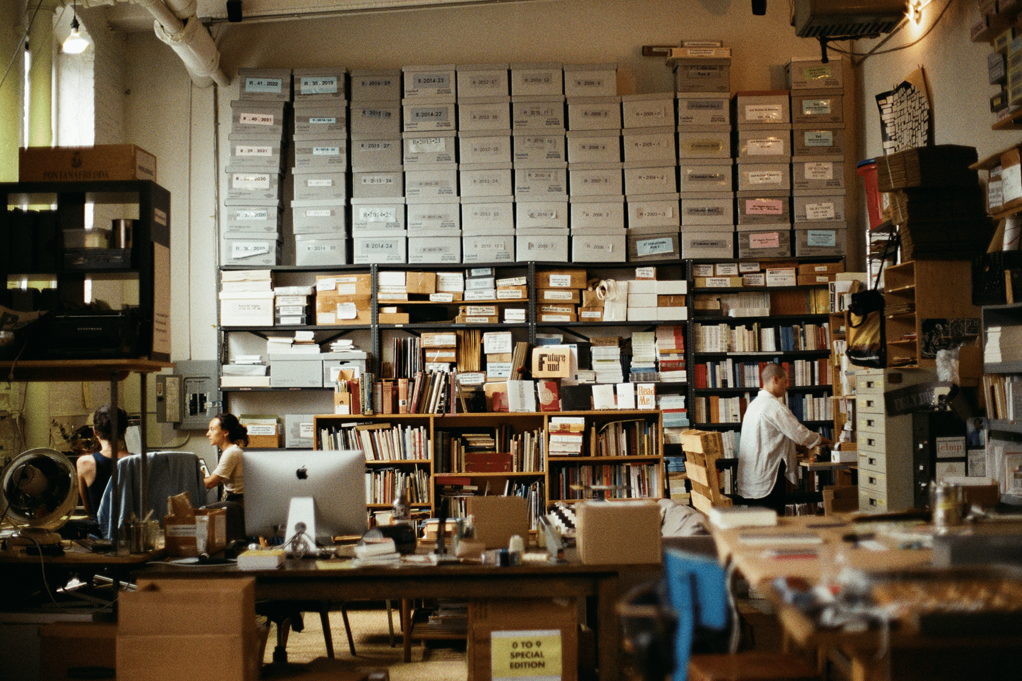 Large room filled with boxes on shelves, desks, and two people at the desks.