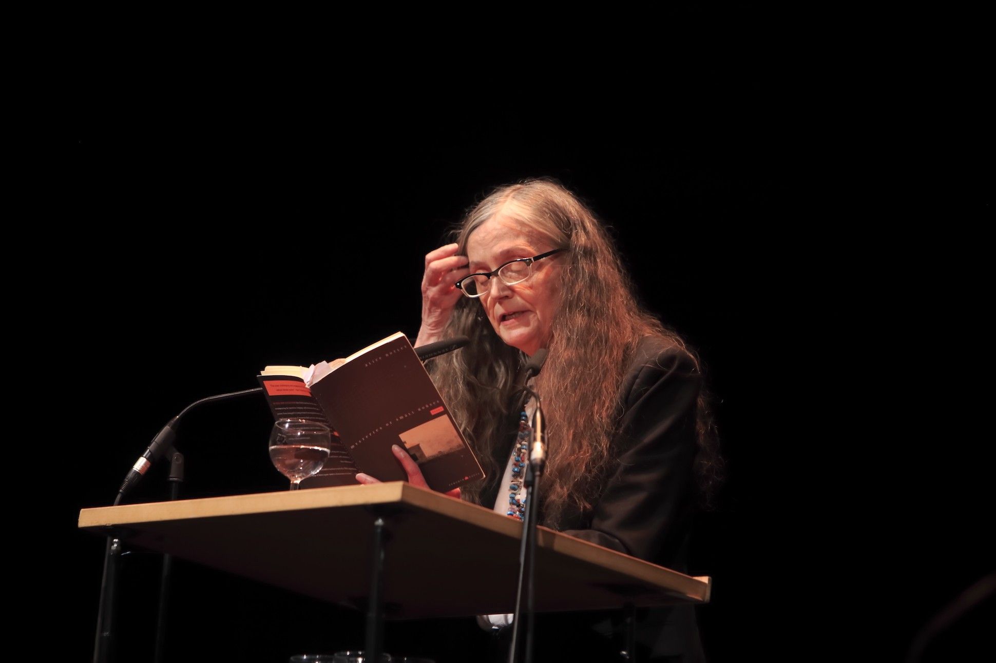 Alice Notley stands at a lectern reading from a book.