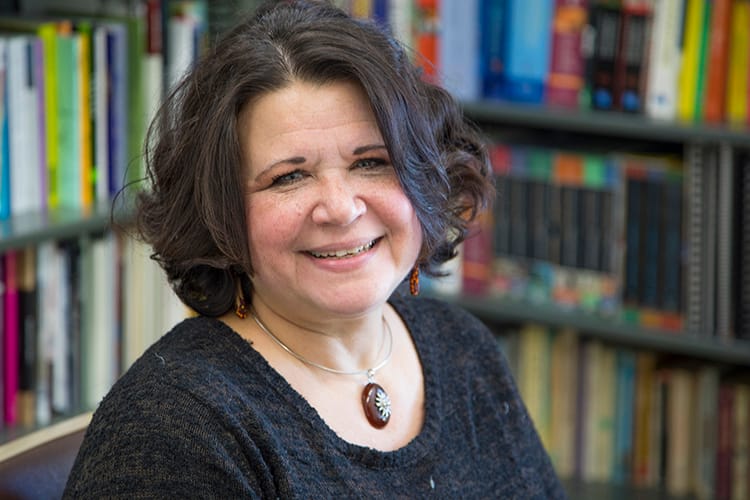 Brenda Cárdenas, a light-skinned Chicana woman with dark brown hair and wearing a brown sweater sits in front of a bookcase.