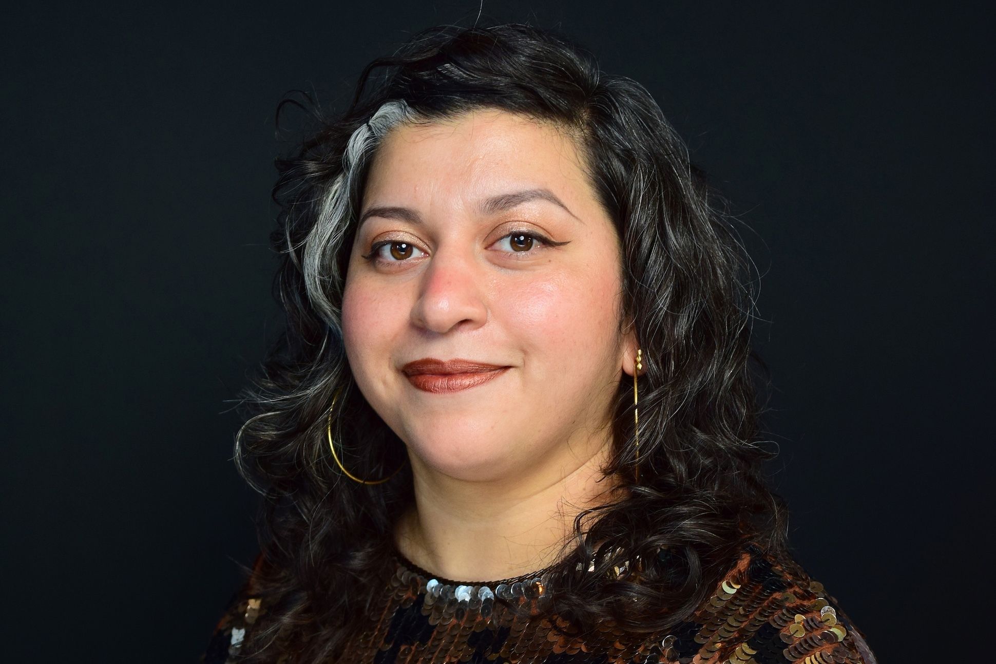 Victoria Martinez, a Latina woman with brown hair and a grey streak, wearing a sparkly top.