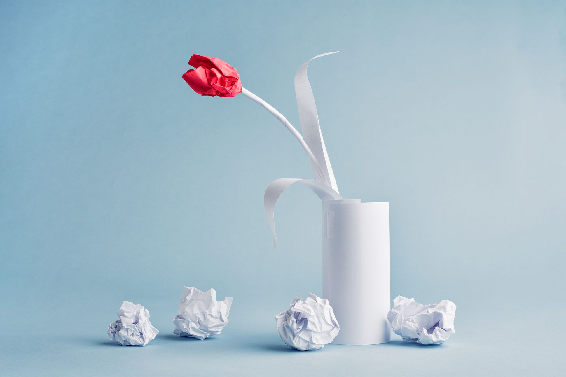 A vase made of paper with a red paper flower is surrounded by crumpled up balls of paper in front of a sky blue background.