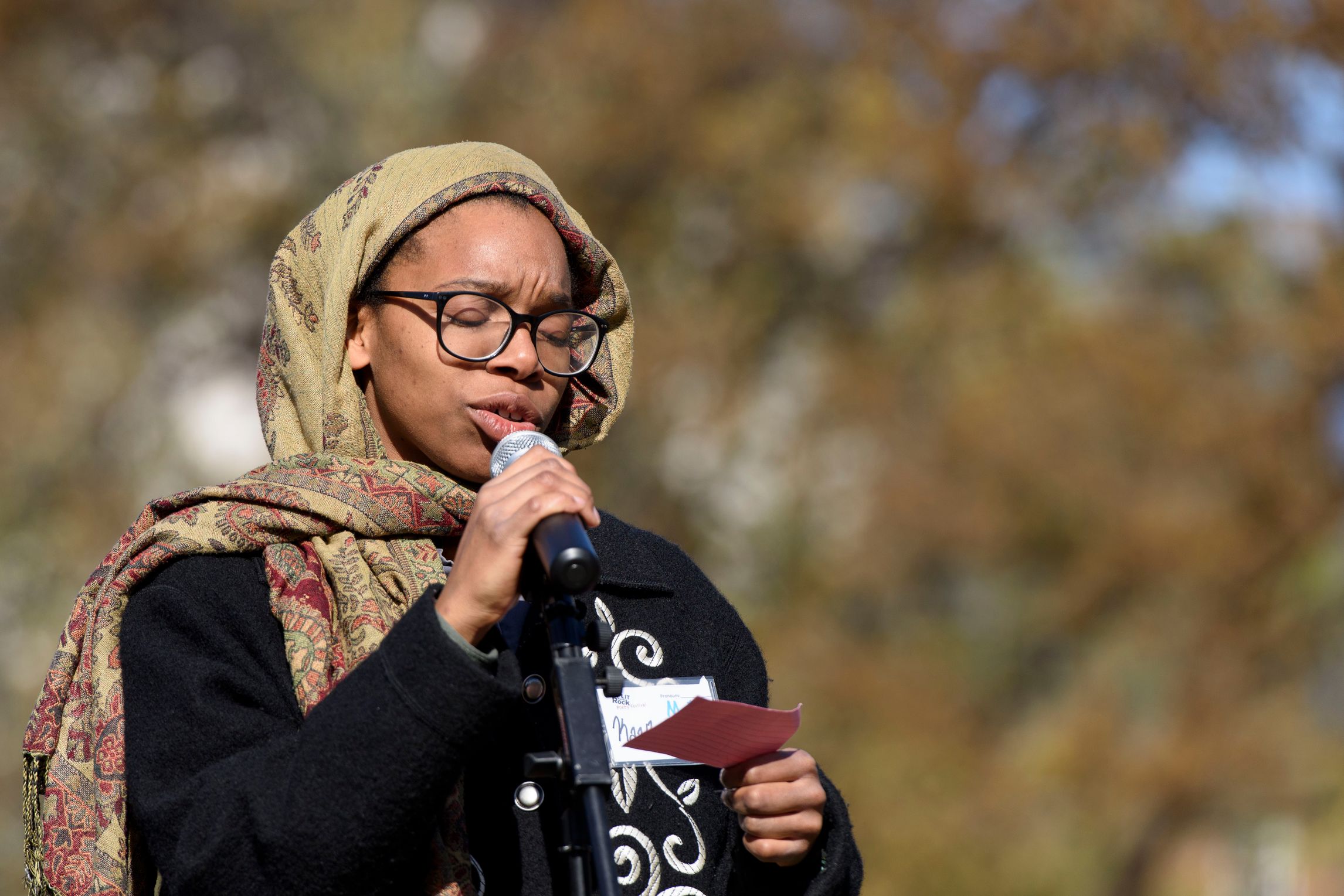 Person speaking into a microphone with their eyes closed, holding a notecard, with blurred trees in the background, glasses, and a headscarf.