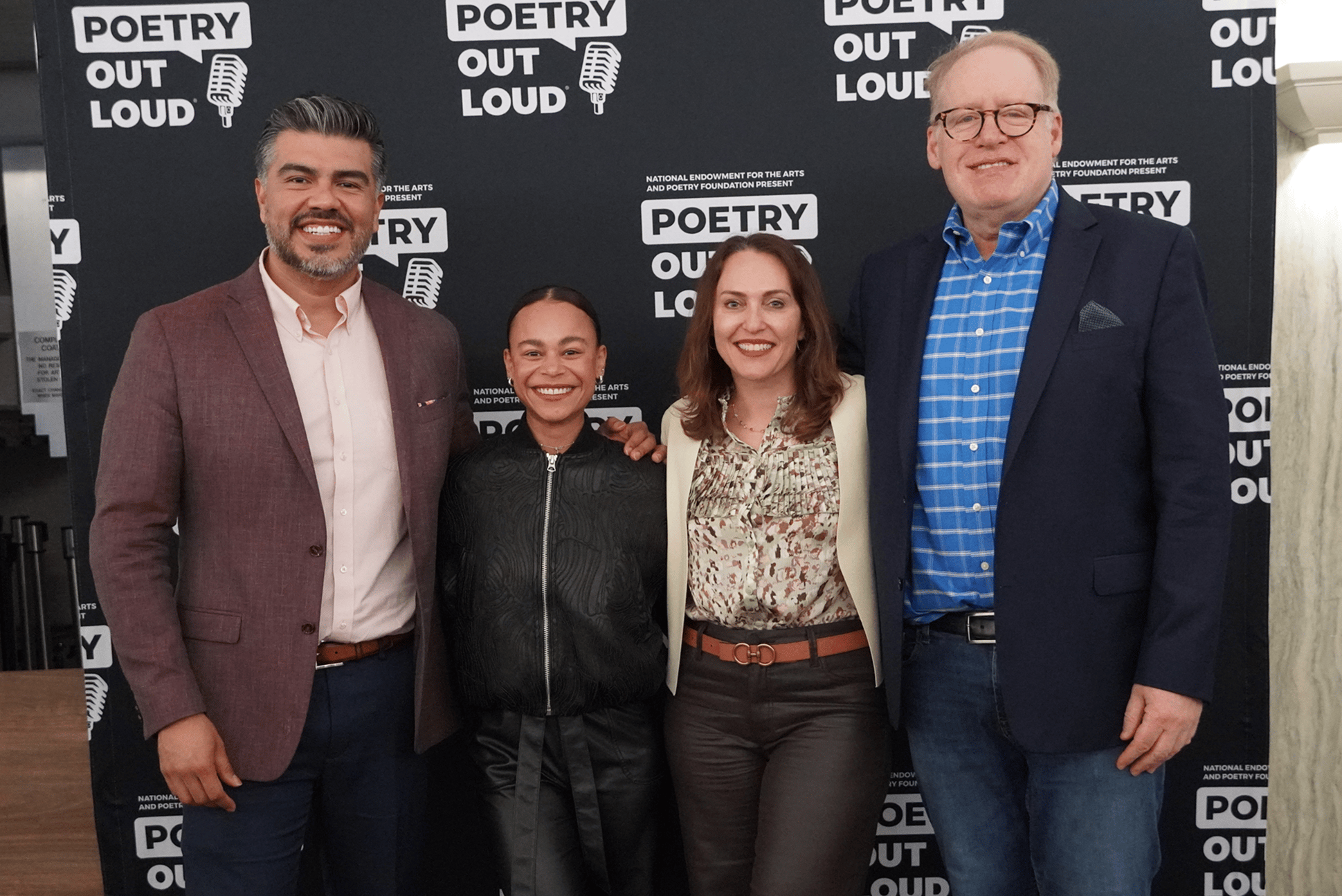 Four adults pose with arms linked in front of a black backdrop with text Poetry Out Loud and an image of a microphone