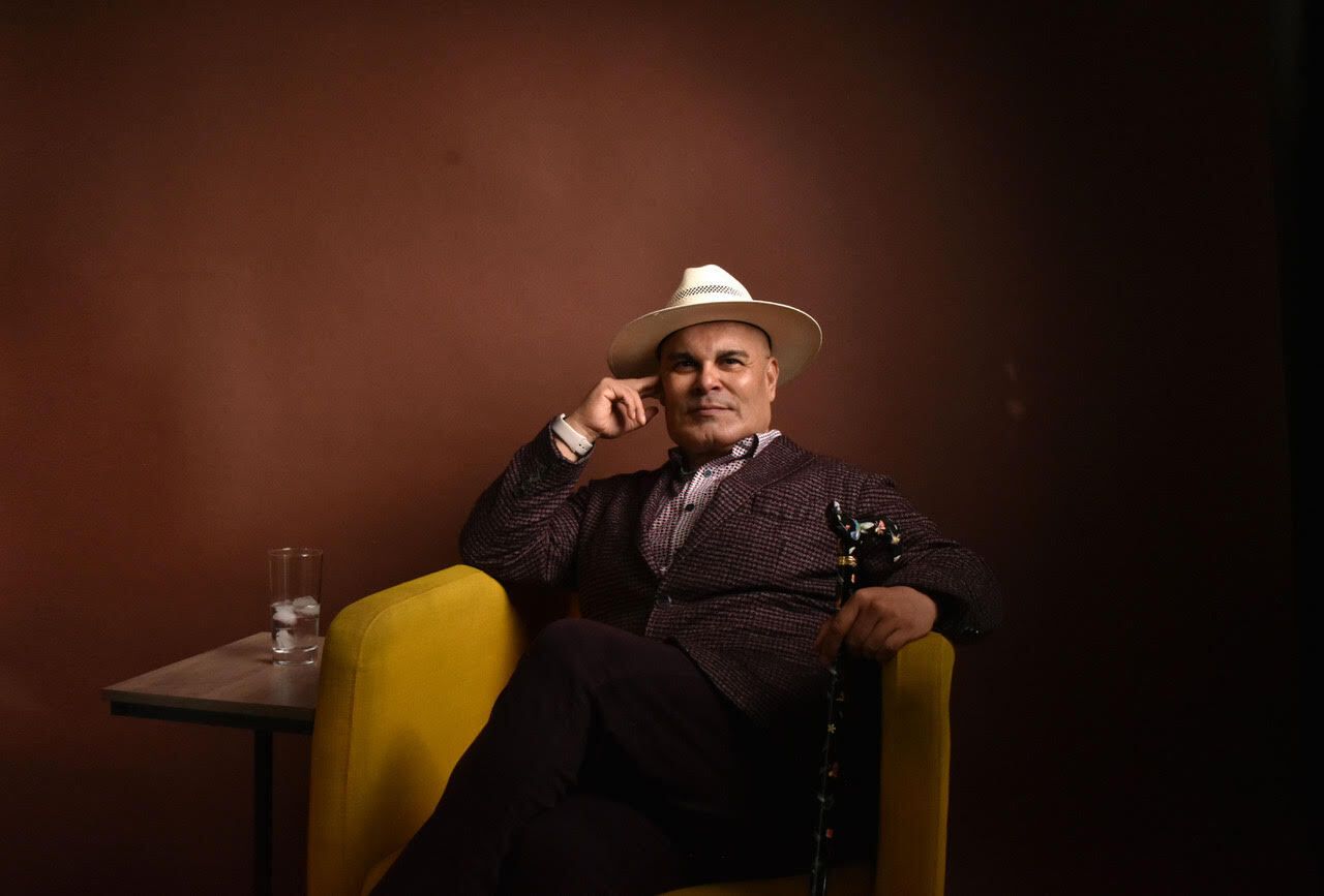 Rigoberto González sits in a yellow chair; he wears a dark suit, light hat, and holds a cane.