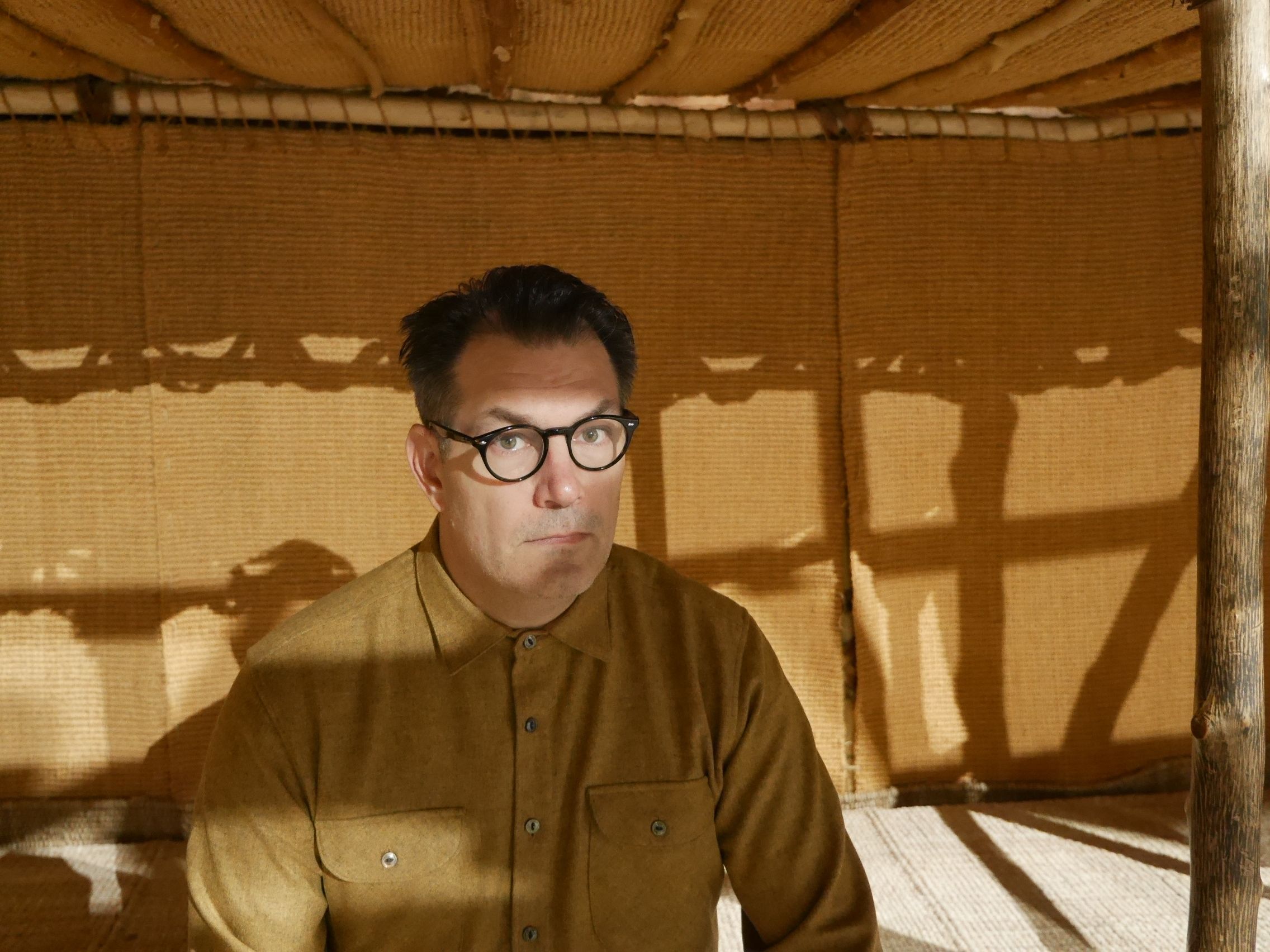 A person in glasses and a button-up shirt sits inside a woven structure, with sunlight and wooden beams casting patterned shadows.