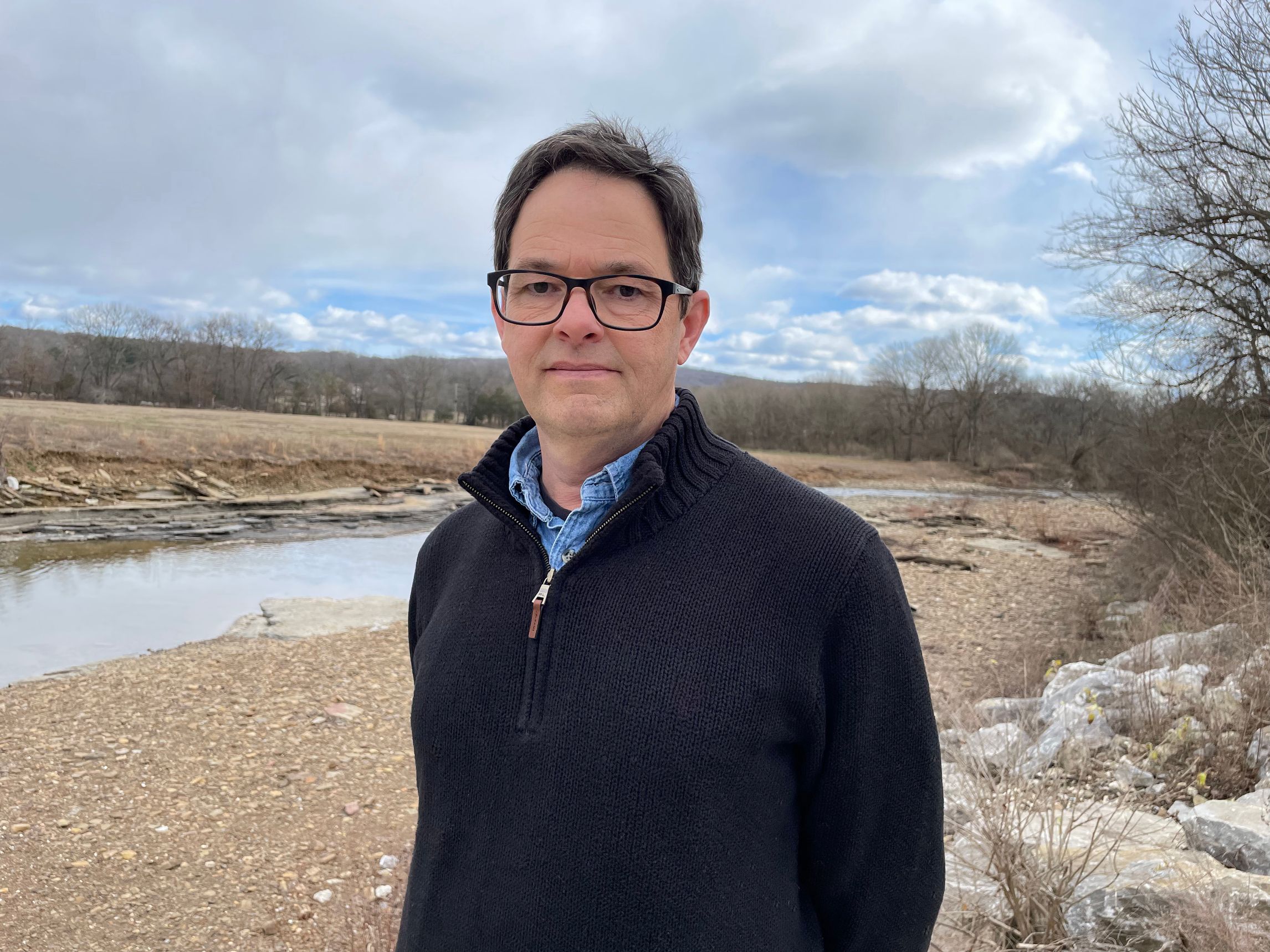 Davis Mc Combs, a spectacled white male in a blue sweater standing outside by a thin stream of river.