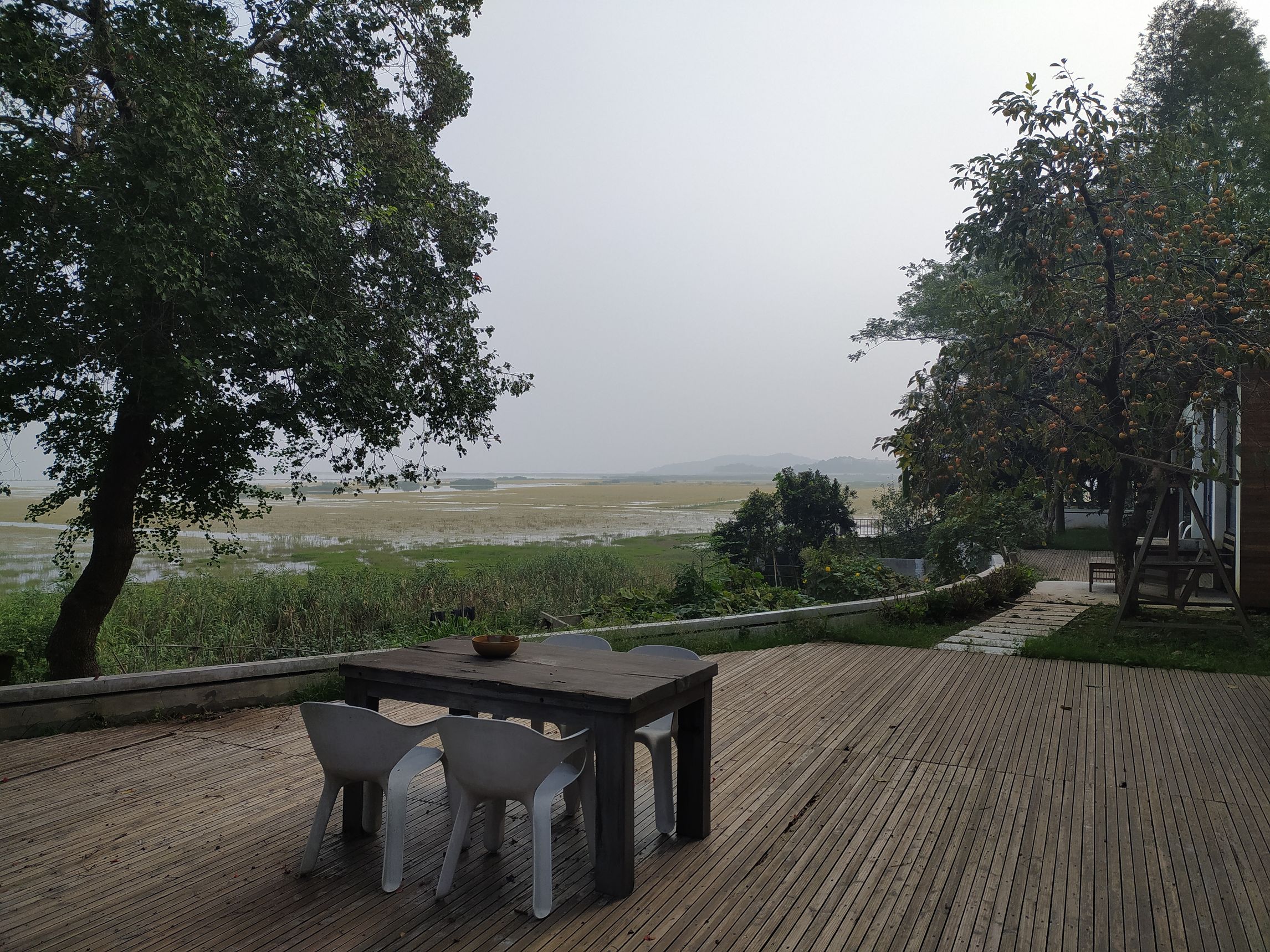 a table and chairs sit on a large wooden deck with fruit trees and fields beyond.
