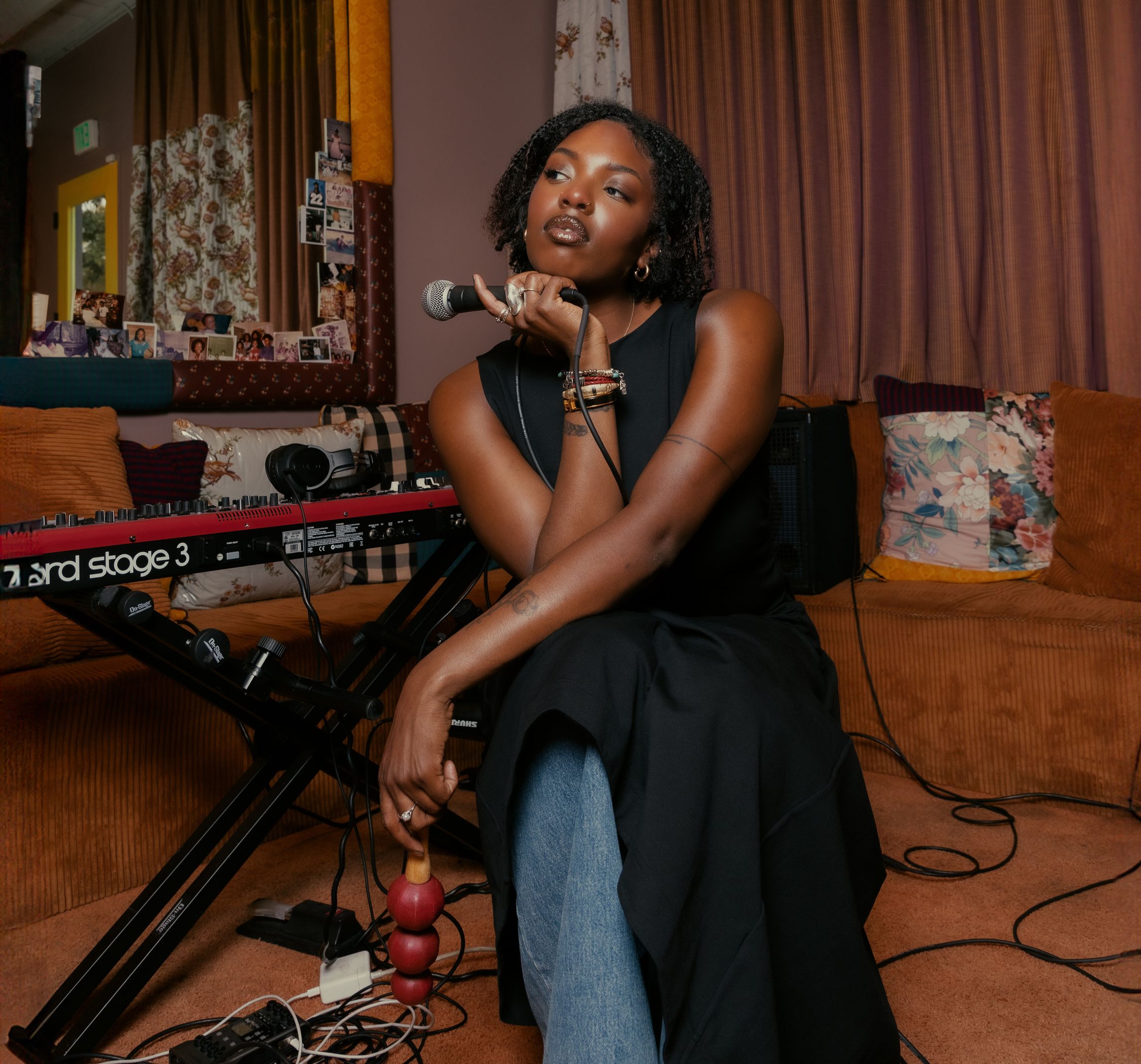 Shawnee Dez, a Black woman with dark brown hair, posed with a Ghanian rattle shaker, in front of a Rhodes keyboard.