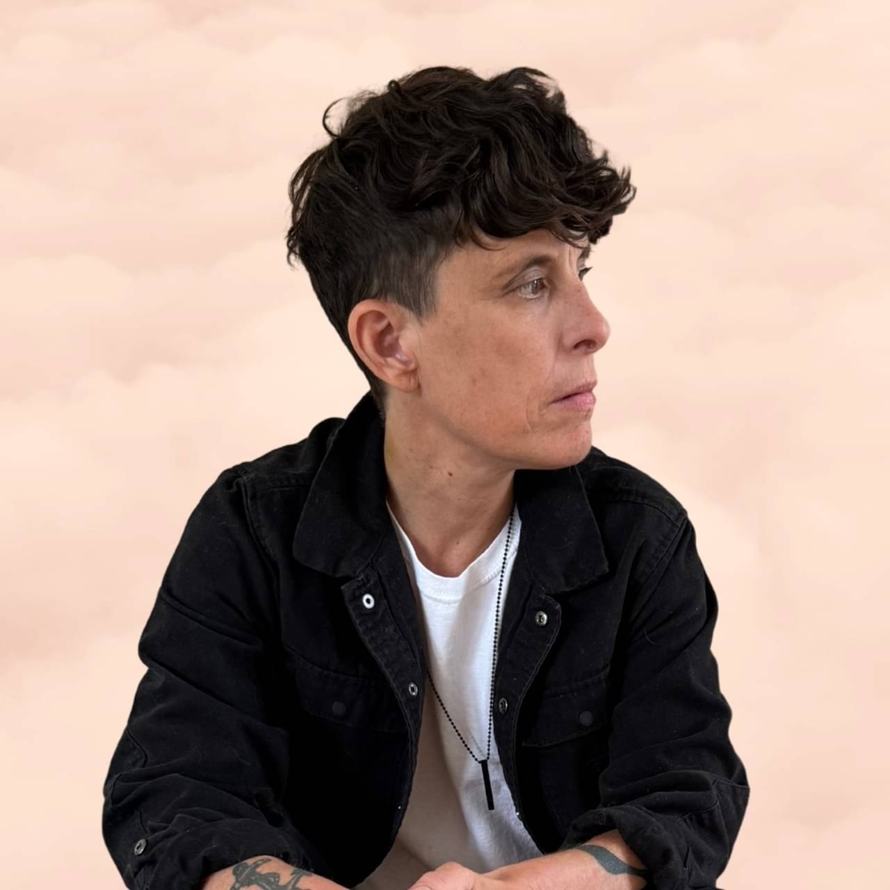 Side profile of Gibson with short hair, wearing a black jacket over a white top, with a silver chain hanging around their neck, tattoos on forearms, against a light pink/peach background.
