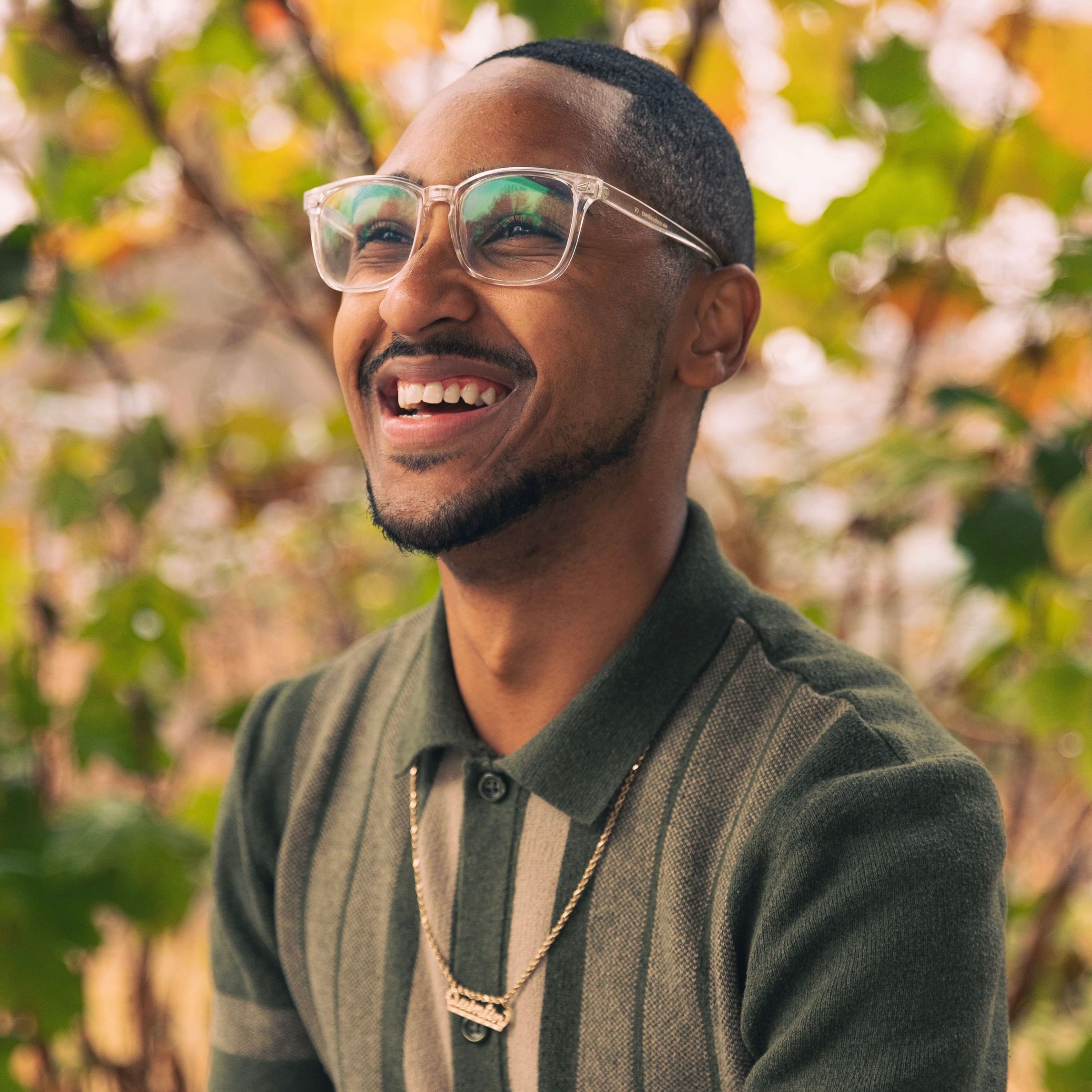 DeeSoul Carson, a black person with short black hair and a green shirt, smiles in front of a background of leaves