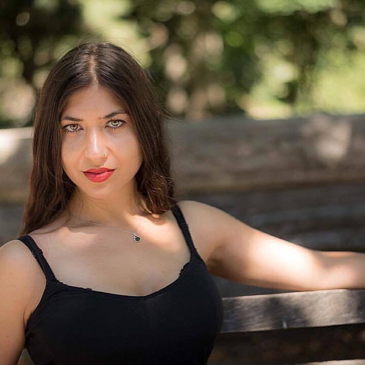 Jezmina Von Thiele, a woman with brown hair, olive skin, and green-hazel eyes, wearing a black tank top, sits outside on a bench with trees in the background