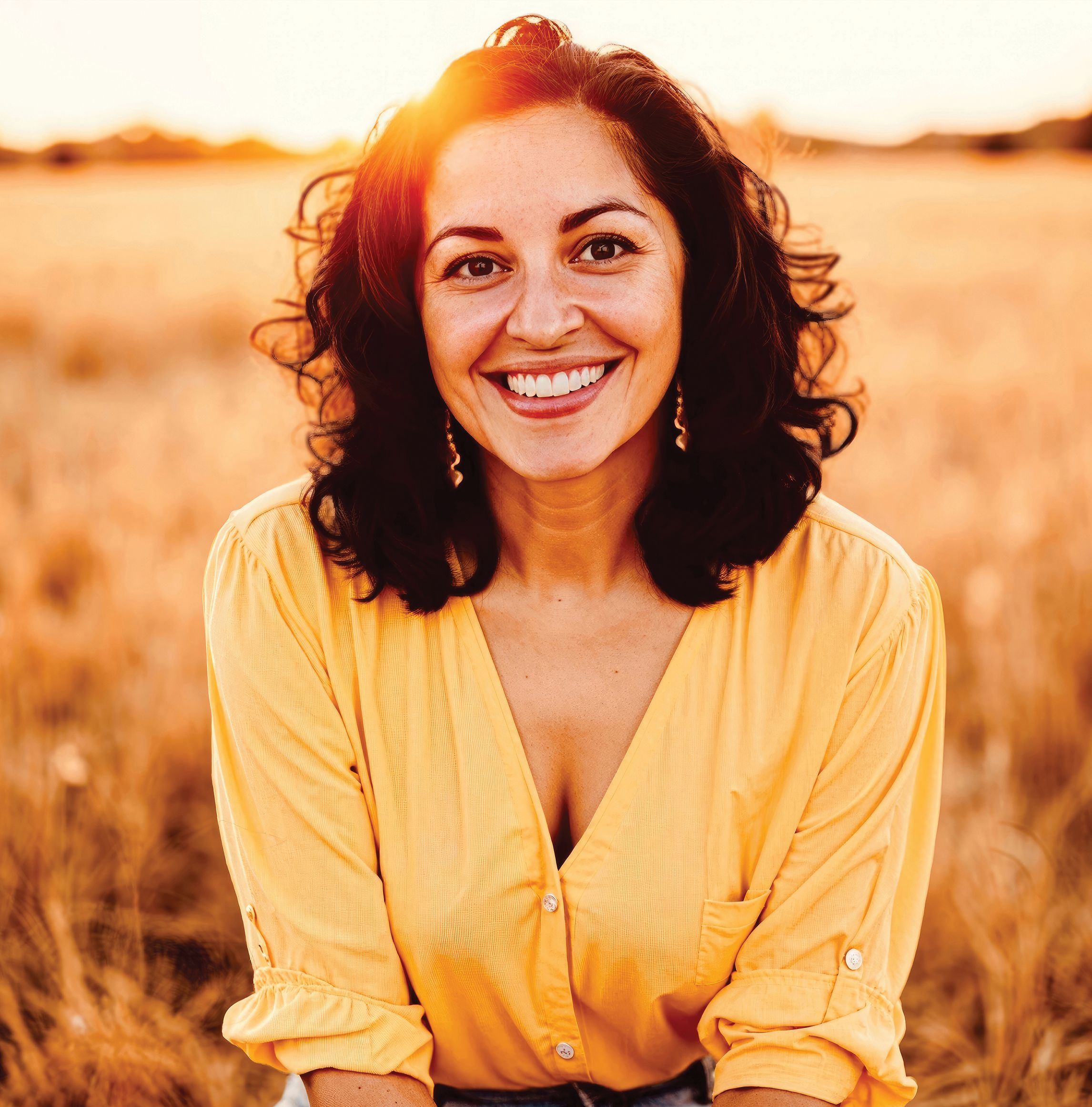 Aimee Nezhukumatathil sits in a field at sunset wearing a yellow shirt and smiling at the camera