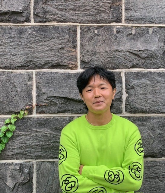 Andrew Chi Keong Yim, an Asian man with black hair, stands in front of a brick wall with his arms crossed, wearing a neon green sweater