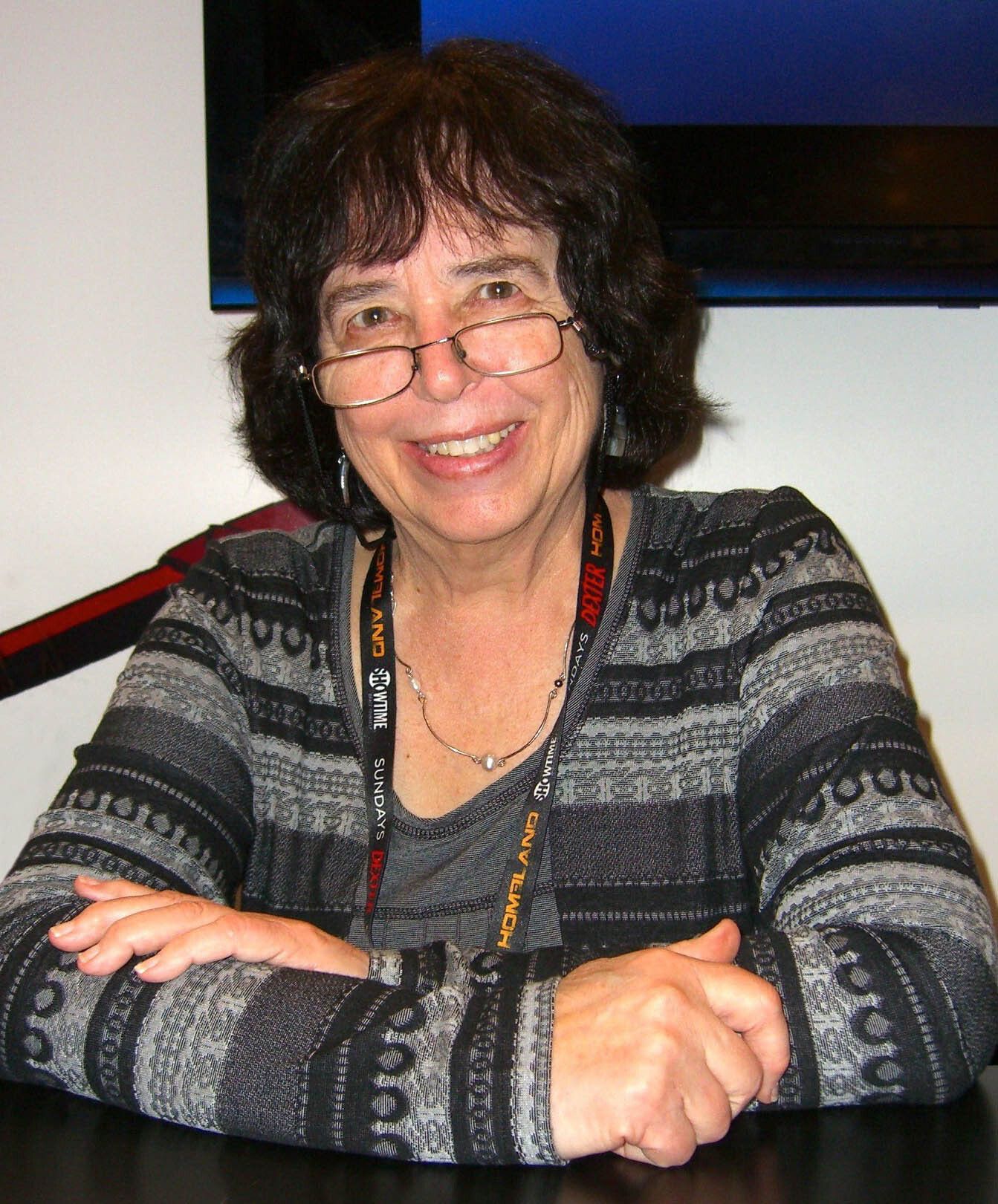 Portrait of Jane Yolen wearing glasses and a striped sweater seated at a table