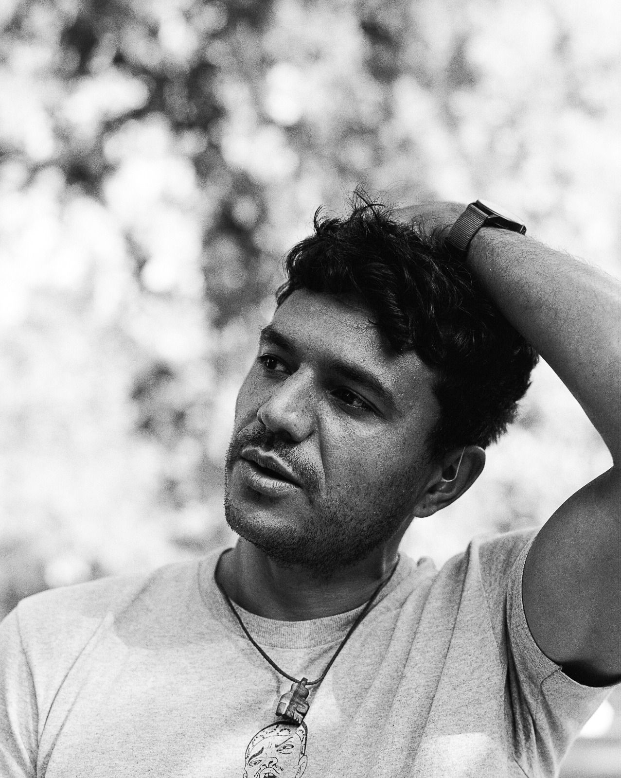 A black and white photo of the poet&#039;s head and shoulders, in a white t-shirt, one hand on his head, looking out of the frame
