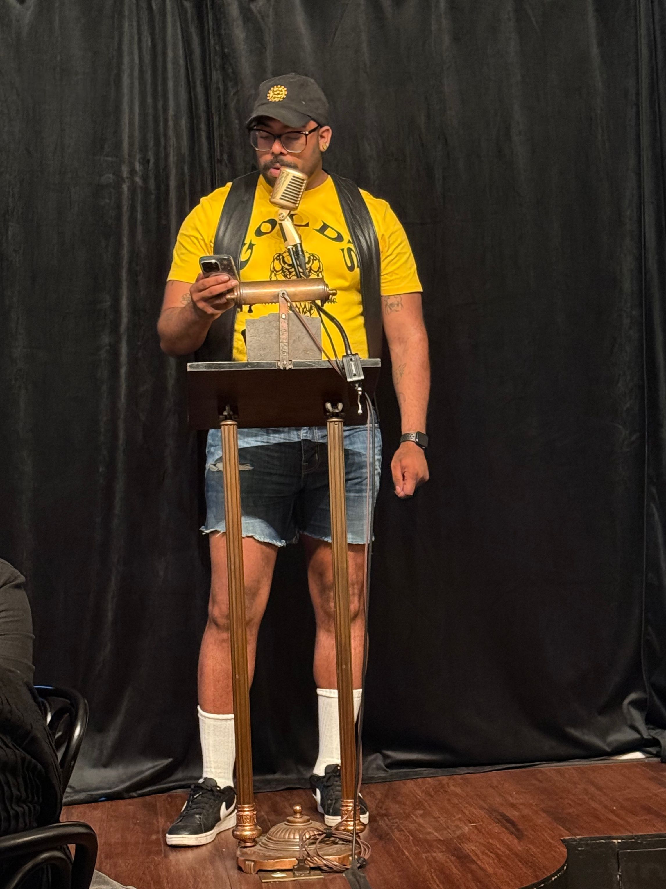 Queer Black Human standing at at podium wearing a Yellow Gold's Shirt and a Jean shorts, also a leather vest and black hat. Behind them there is a Brooklyn Poets banner.
