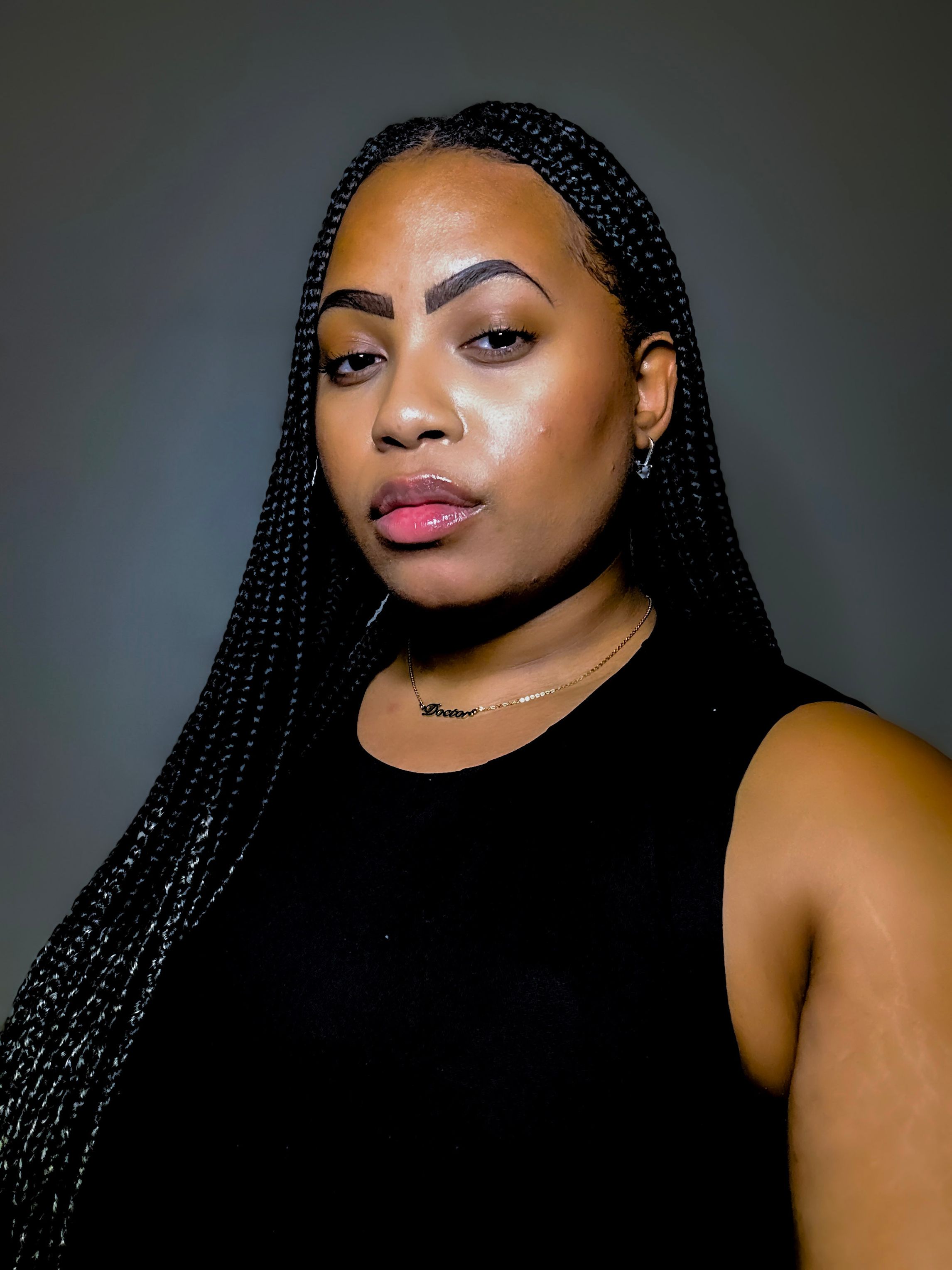 Taylor Byas, a Black woman with black braids, wears a black, sleeveless shirt and gold necklace, stands in front of a grey background.