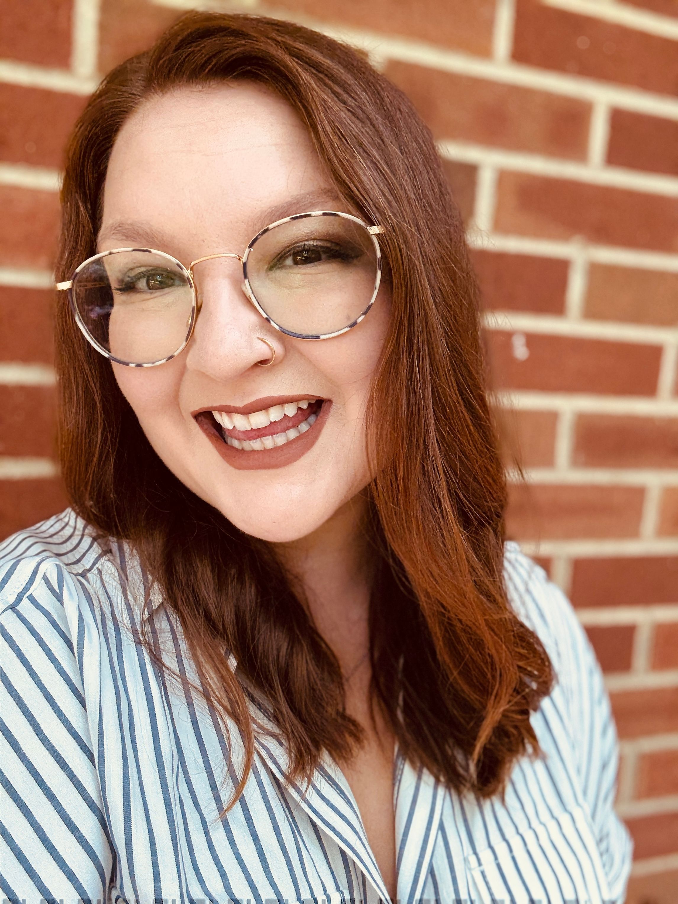 Hannah V Warren, a white woman with red hair, smiles in front of a brick wall.