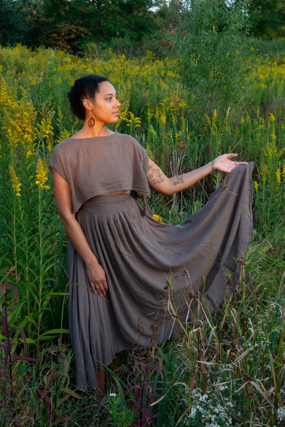 A.Martinez, a woman with brown skin and black short curly hair braided in the front, holds her grey skirt out to the side with her left hand, looking towards it, and standing in a field of goldenrods in late Summer golden hour