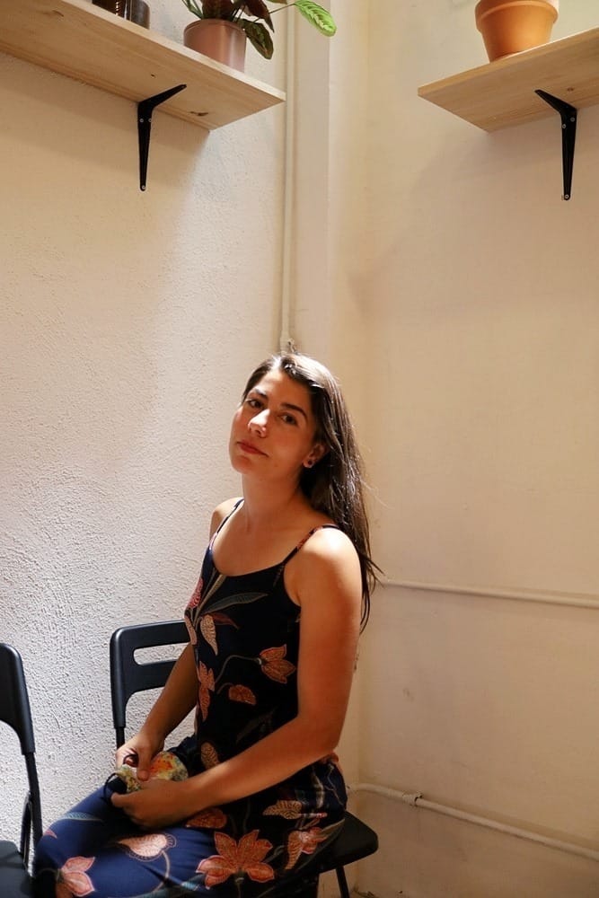 Ruth Llana, olive skin woman with long brown hair, wears a blue dress with flowers, and sits on a black chair in front of a wall, with the light cascading on her shoulders and face.
