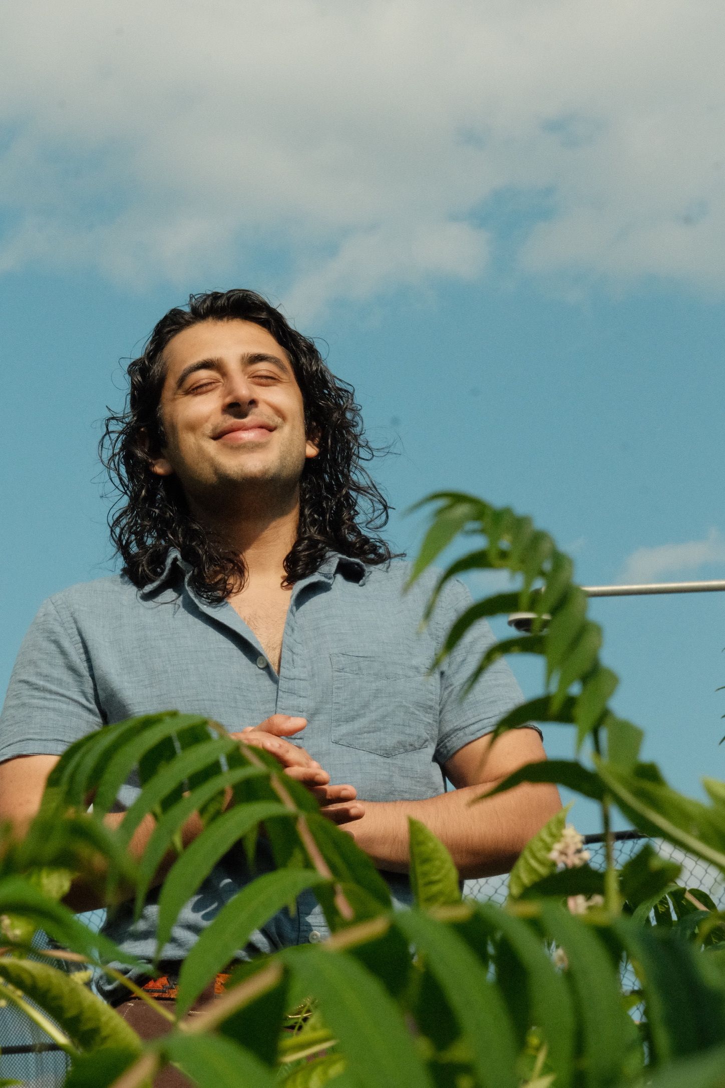 Andres Cordoba, a Colombian-Peruvian man with shoulder-length wavy, black hair stands in a blue-collared shirt with his hands clasped in front of him. Looking towards the sun, his eyes are closed and he is smiling. The photo is from a lower-angle and behind him is a slightly-clouded blue sky. In the foreground, covering his lower-half, there is foliage.