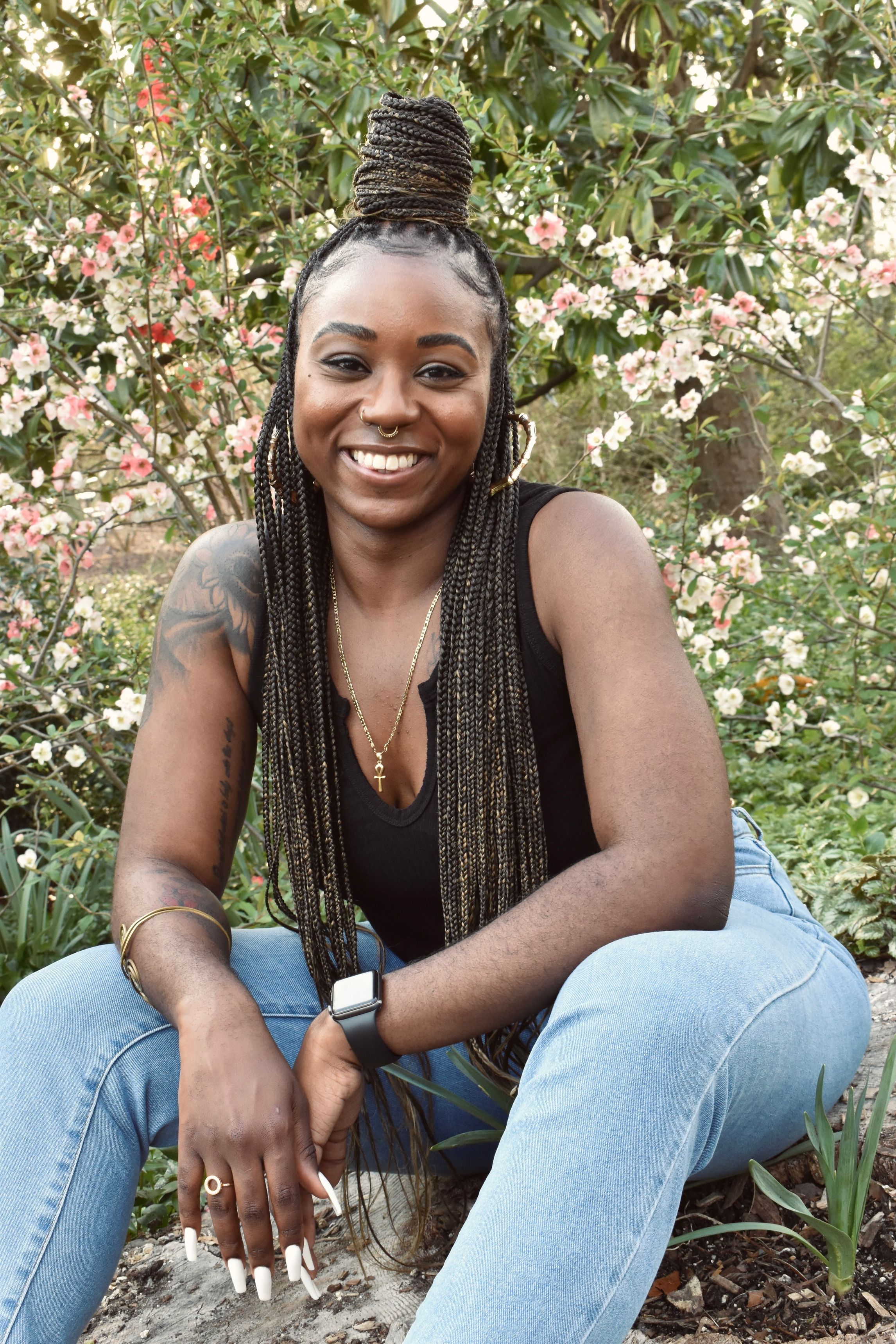 Shaina Phenix, Black woman with black box braids and blonde highlights. Black shirt, blue jeans. In front of white and pink flowers.