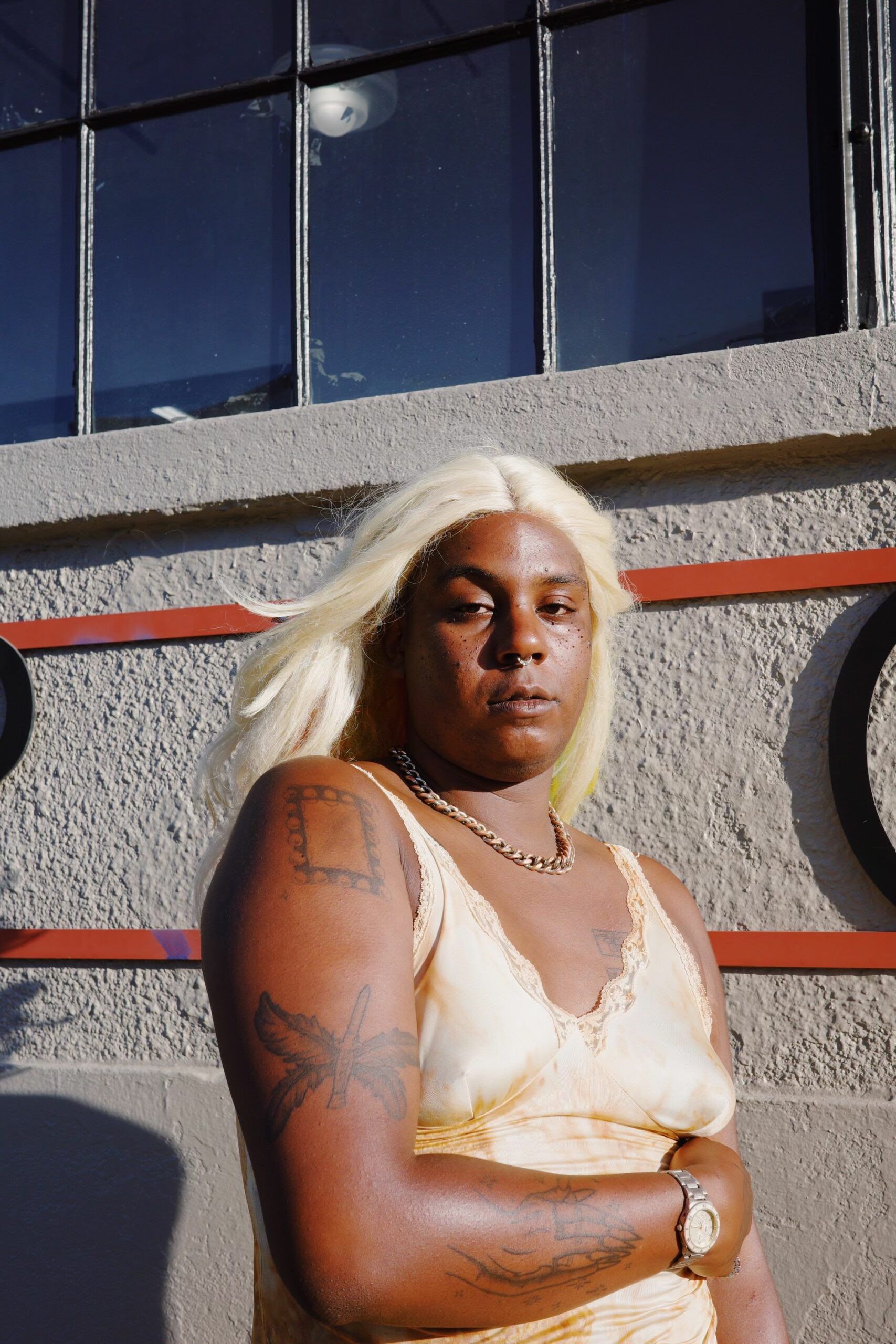 Jzl Jmz, a Black woman with blonde hair, crossing her arms, wearing a rust-bleached lace & satin gown, in front of a grey wall with windows & decals