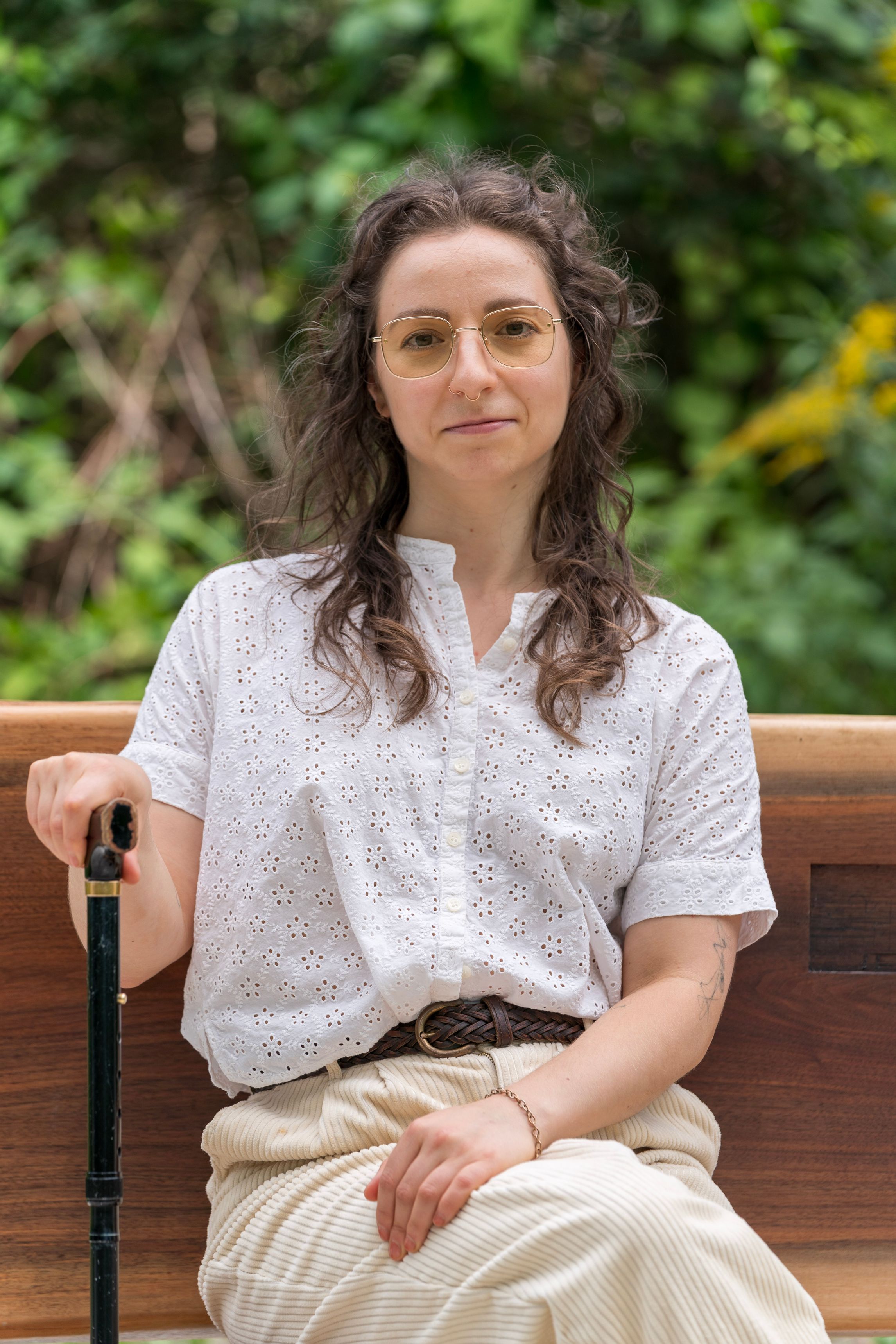 Leigh is a White woman, smiling slightly, with wavy, shoulder-length brown hair. She wears gold-rimmed glasses, a whLeigh is a White woman, smiling slightly, with wavy, shoulder-length brown hair. She wears gold-rimmed glasses, a white button-down t-shirt, and tan pants. She sits on a wooden bench, her hand wrapped around the handle of a cane. Greenery is blurred out in the background.
ite button-down t-shirt, and tan pants. She sits on a wooden bench, her hand wrapped around the handle of a cane. Greenery is blurred out in the background.